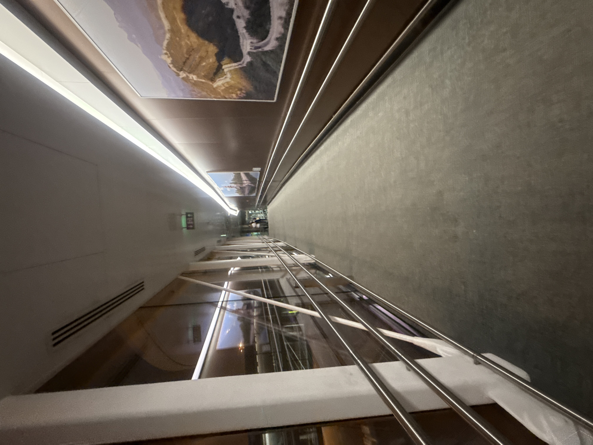 The image shows an interior view of an airport jet bridge, a walkway that connects an airport terminal to an aircraft. The jet bridge is enclosed and features a curved ceiling with integrated lighting. The walls are primarily a dark, warm wood-like material, adorned with two large framed pictures. The picture on the right depicts a section of the Great Wall of China, while the one on the left shows a historical building or landmark.
A polished metal handrail runs along the right side of the walkway, positioned at a lower height for accessibility. The floor is covered in a dark, textured carpet. On the left side of the jet bridge, large windows offer a view of what appears to be an airport tarmac at night, with lights and some indistinct structures visible. A white pillar supports the structure on the far left.
In the distance, at the end of the jet bridge, several individuals are visible, suggesting movement and activity within the airport. A green exit sign with figures is also noticeable on the ceiling in the distance. The overall scene is one of a functional and modern airport transit area. The provided image does not offer any specific visual cues to pinpoint the location within Guangyang District, China.
