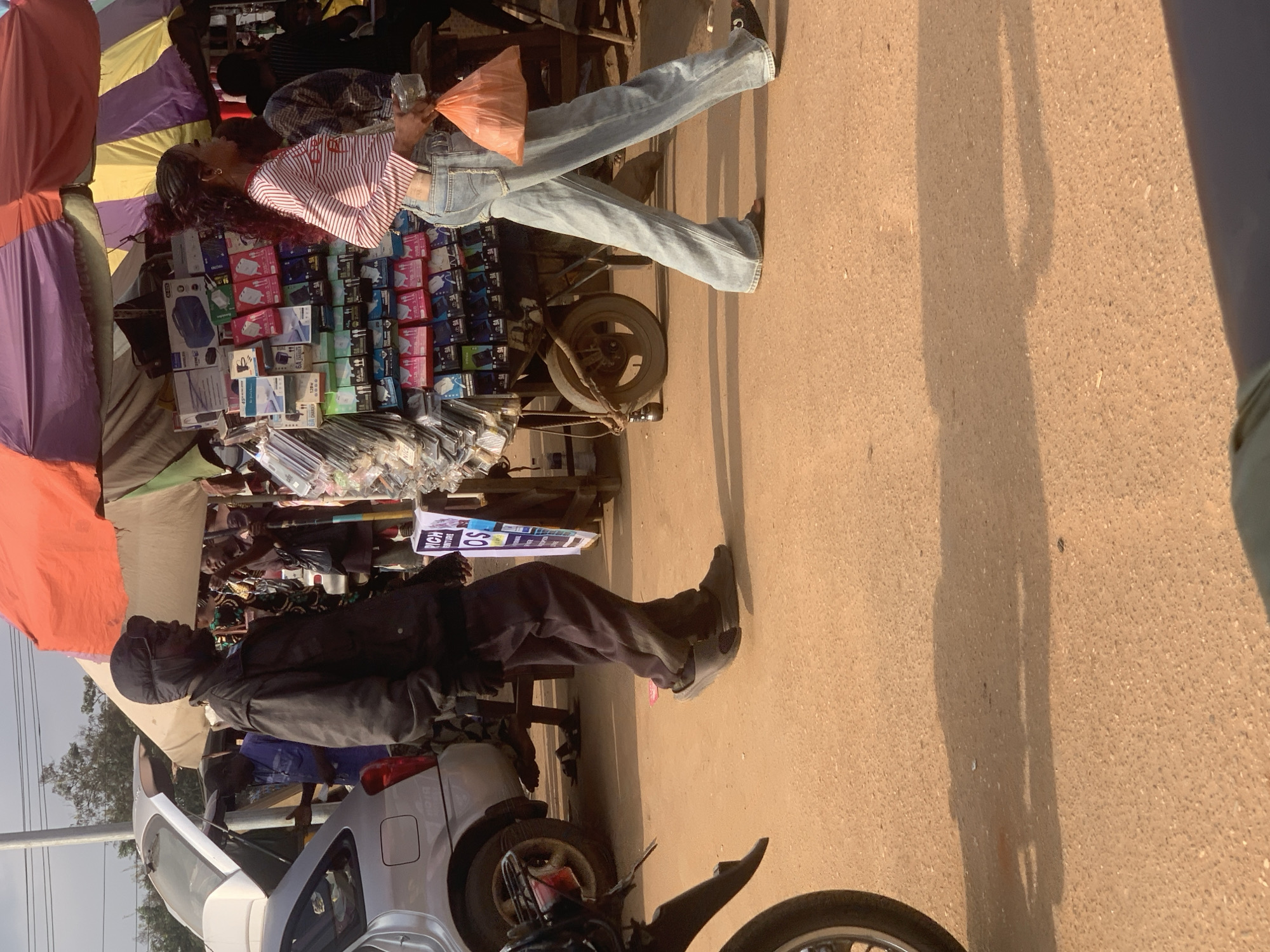 The image captures a street scene in Shagamu, Nigeria, near a market stall. In the foreground, a person in dark clothing and a cap is walking away from the camera. To their right, a woman with long, dark hair, wearing a striped shirt and blue jeans, is also walking away, holding an orange plastic bag and a transparent container. Behind them, a mobile market stall displays various electronic accessories. Further back, a silver car with its trunk open is parked on the left. The ground is a dirt or gravel road, and the background shows more market stalls and trees under a bright sky.