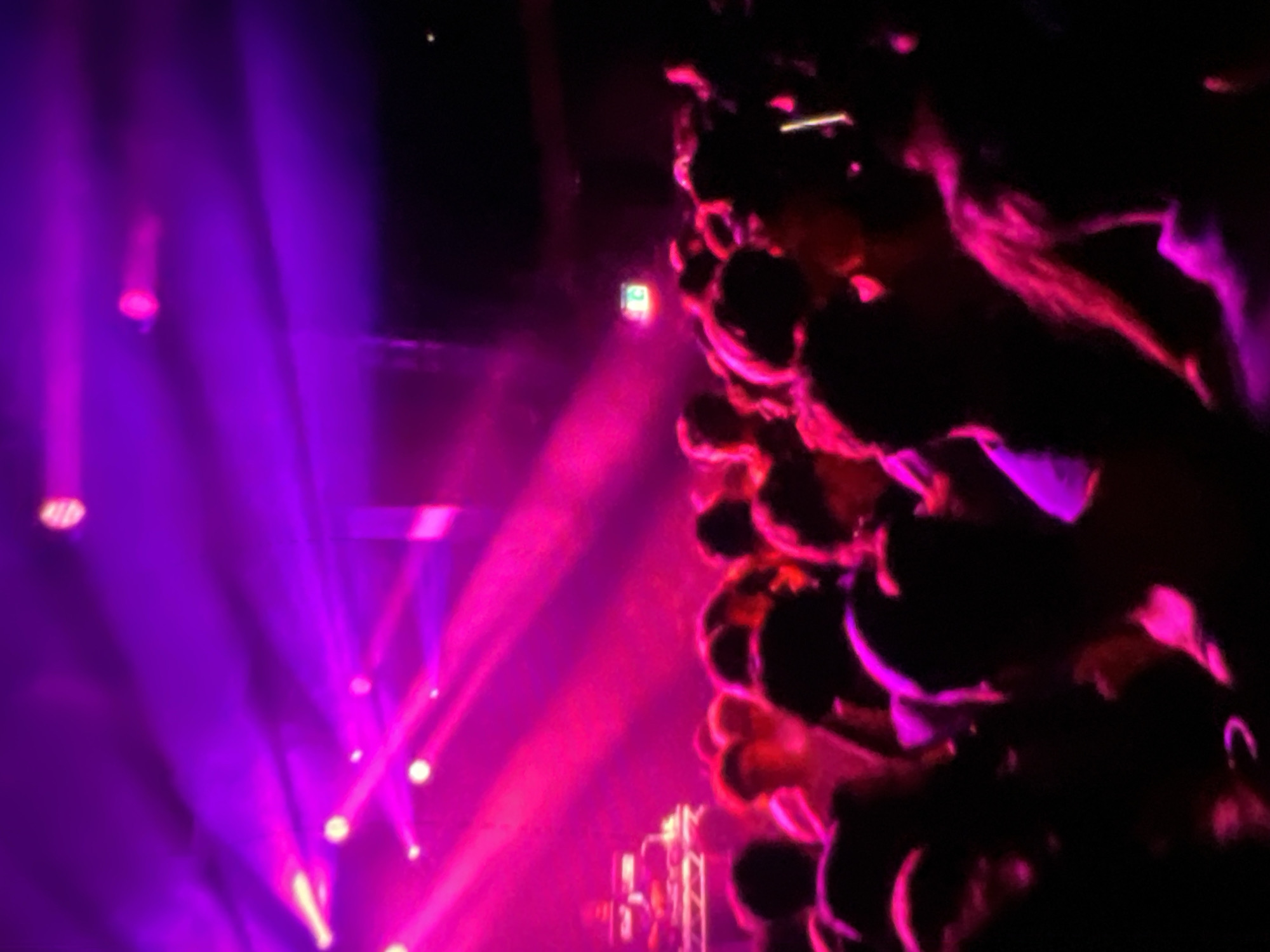 The image depicts a dimly lit indoor venue, likely a nightclub or concert hall, illuminated by vibrant purple and pink stage lighting. Beams of light crisscross the smoky atmosphere, creating a dramatic and energetic ambiance. In the foreground and midground, a crowd of people are visible, their forms silhouetted against the colorful lights. Some individuals appear to be dancing or engaged in conversation, their faces obscured by the low light and the density of the crowd. In the background, a DJ booth or stage area with electronic equipment is faintly discernible, suggesting a performance or musical event. The overall impression is one of a lively social gathering taking place in Lagos, Nigeria, characterized by its dynamic lighting and palpable sense of energy.