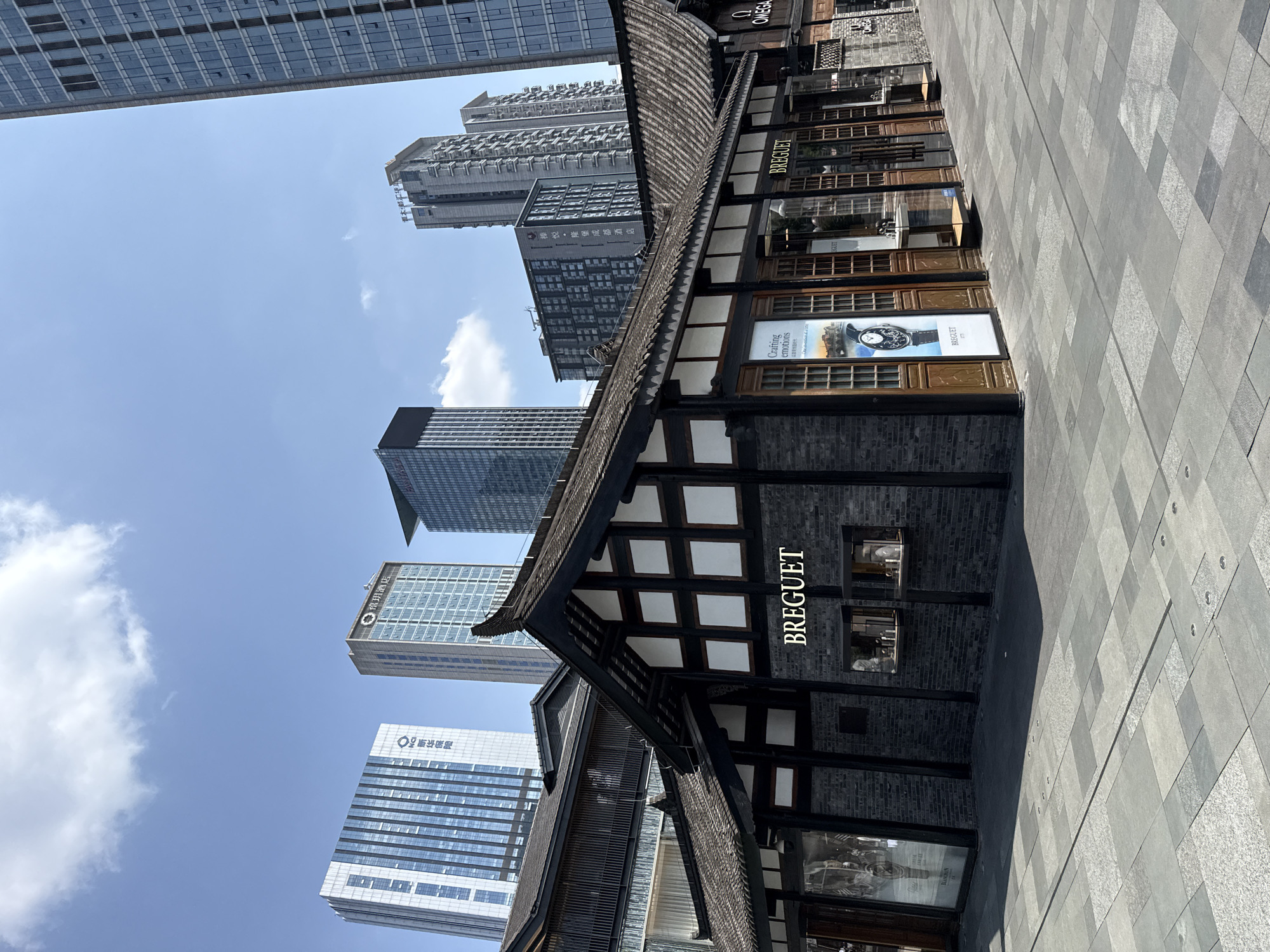 The image captures a street scene in Jinjiang District, China. In the foreground, a traditional-style dark grey brick building with white squares bears the "BREGUET" logo, indicating a luxury watch store. Its eaves extend outward, sheltering windows displaying watch advertisements. To the right, another sign reads "OMEGA." The foreground is dominated by a grey paved pedestrian walkway with a geometric pattern.
In the background, modern skyscrapers loom, juxtaposing the traditional building with the contemporary urban landscape. A building labeled "新华保险" (Xinhua Insurance) and "榛悦・隆堡成都酒店" (Brandlin Suites Chengdu) are visible. The sky is blue with scattered clouds. The angle of the photograph suggests it was taken from a low vantage point looking upward, highlighting the height of the surrounding buildings.