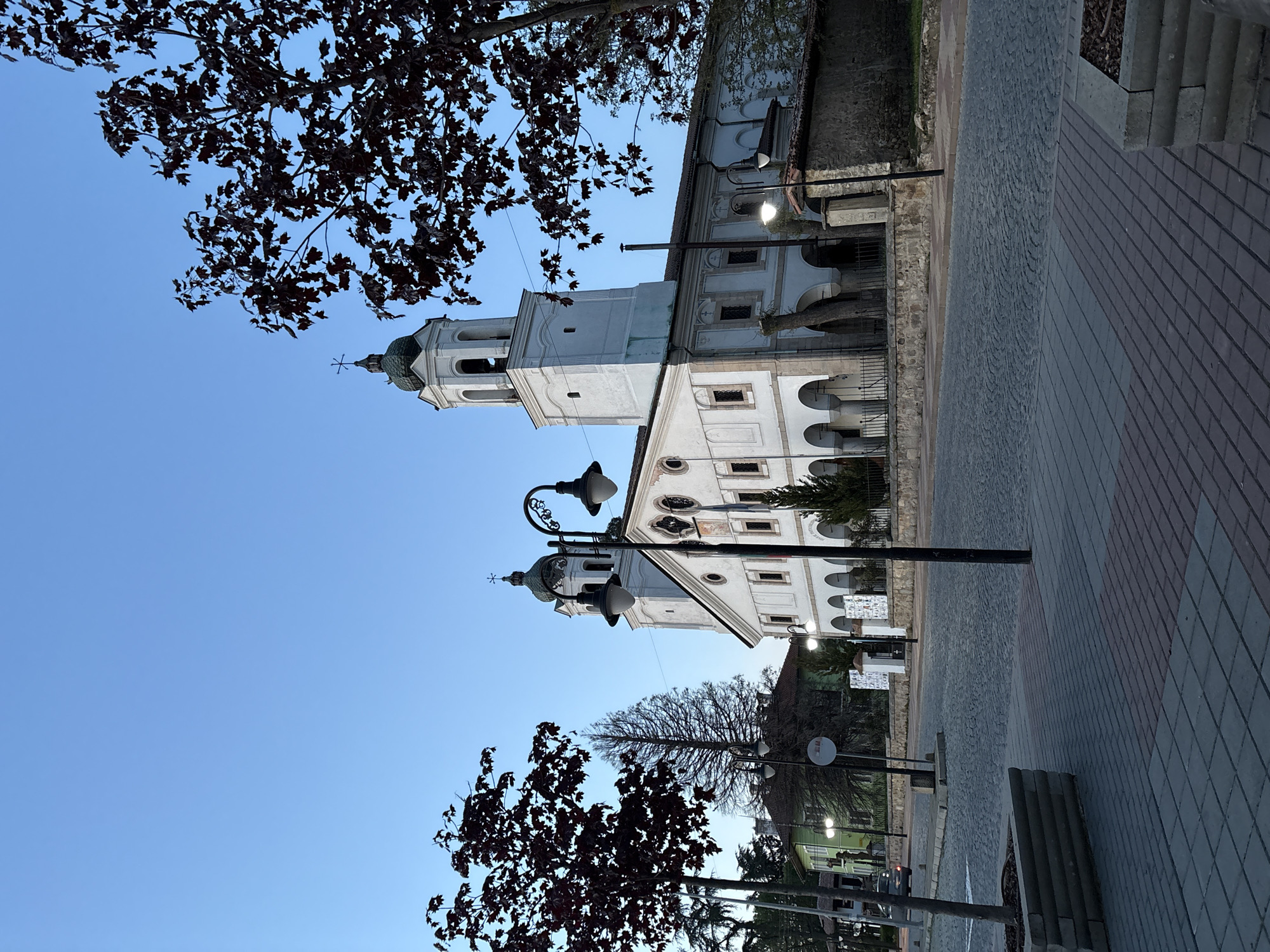 The image shows the Assumption Church in Panagyurishte, Bulgaria. The church is a white, multi-story building with a tall bell tower topped with a cross. A street with a paved walkway runs along the side of the church. There are street lamps and trees in the foreground. The sky is blue.