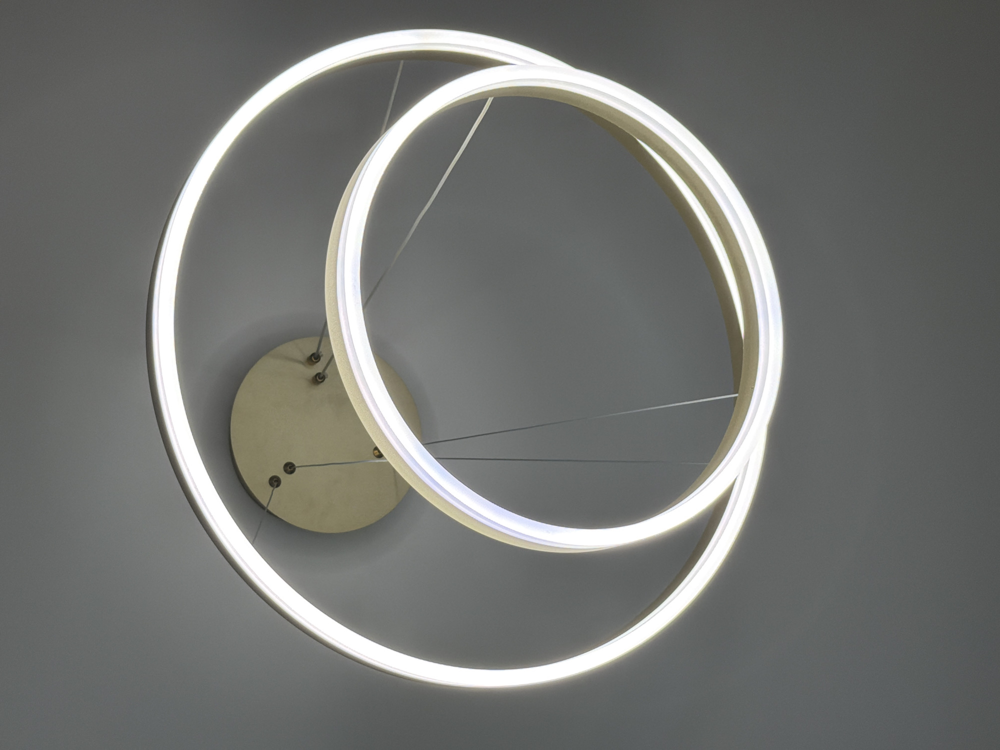 The image shows a modern ceiling light fixture consisting of two intersecting illuminated rings. The rings are suspended by thin cables from a circular mounting plate, which is attached to a gray or off-white ceiling. There are no people or other objects visible in the frame. The environment appears to be a room with neutral-colored walls, likely indoors. There is no information suggesting the location is Moscow, Russia.