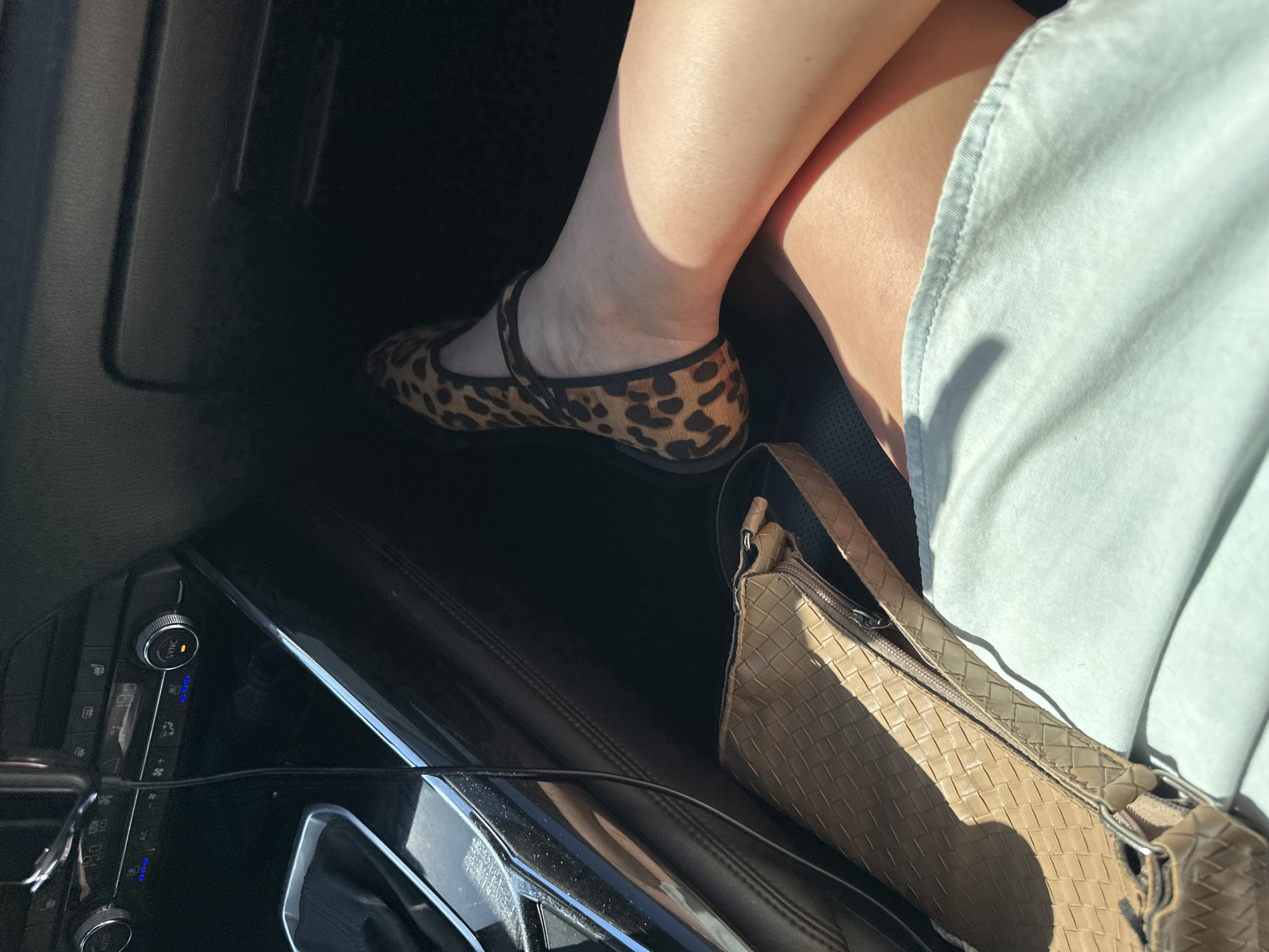 The image appears to be taken inside a car. A person is wearing a light-blue dress. The individual has on a pair of leopard-print shoes. A light-brown handbag with a woven pattern is also visible. The car's dashboard and air conditioning controls can be seen in the foreground. The location cannot be determined from the image alone.