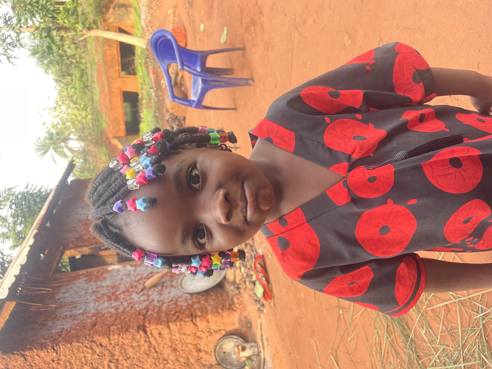 A young girl stands outdoors in Ogbe, Nigeria, wearing a black and red patterned dress and hair adorned with colorful star-shaped beads. A faded blue plastic chair sits behind her near a dilapidated brick building with a corrugated metal roof. The ground is reddish-brown, suggesting a clay-rich soil. The environment has sparse patches of grass.