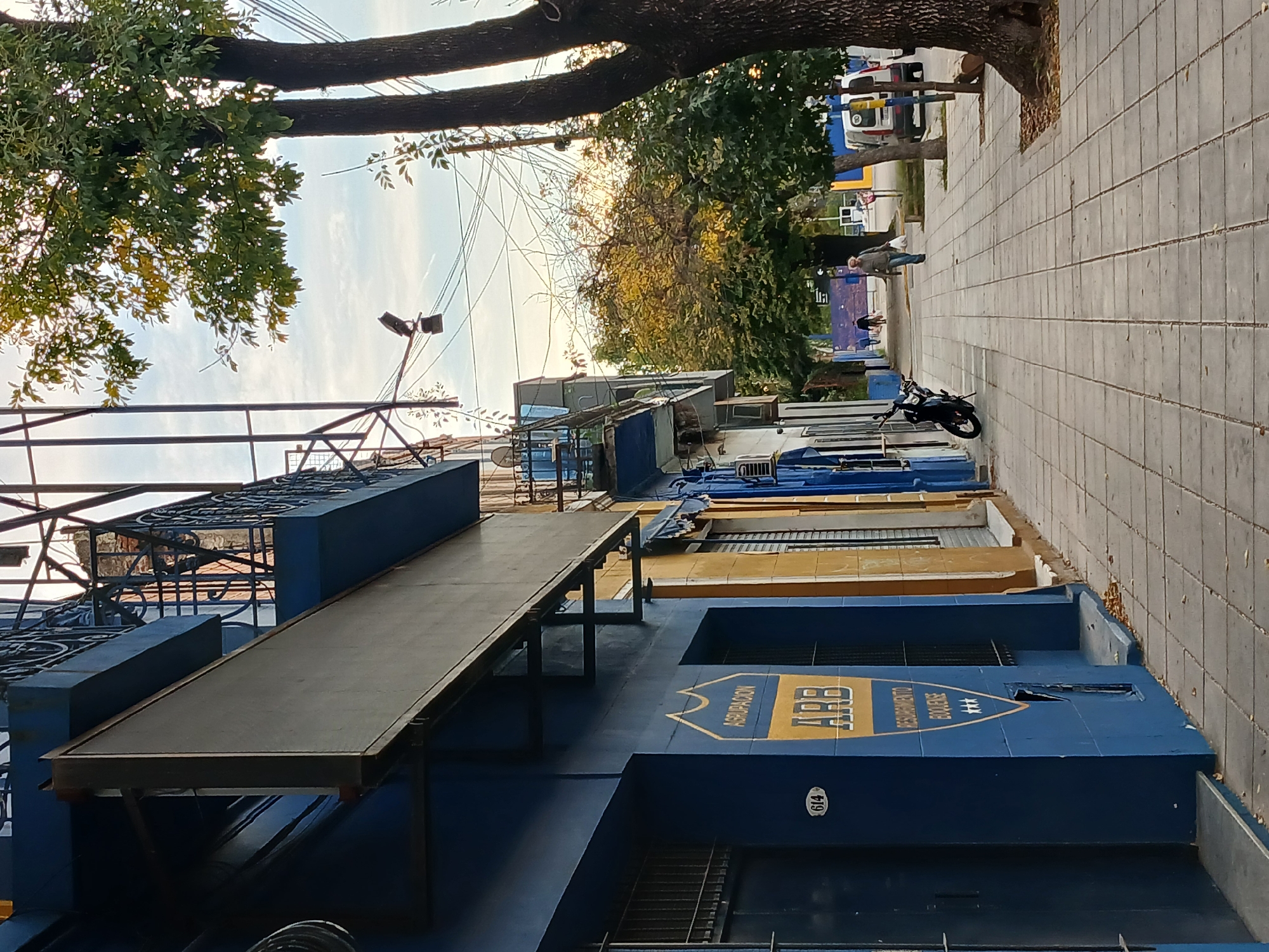 The image shows a street scene in Buenos Aires, Argentina. A blue building with the sign "ASRBU Rescatamento Balerios" is prominent in the foreground, with the number 614 visible on its exterior. A dark-colored motorcycle is parked on the sidewalk in front of a series of buildings, some painted blue and yellow. A person is walking along the sidewalk in the distance, along with white objects in the street. The sidewalk is tiled in gray. A tree is present along the sidewalk, with branches extending into the sky. Overhead are electrical wires and a streetlight. The sky is visible between the buildings and trees.