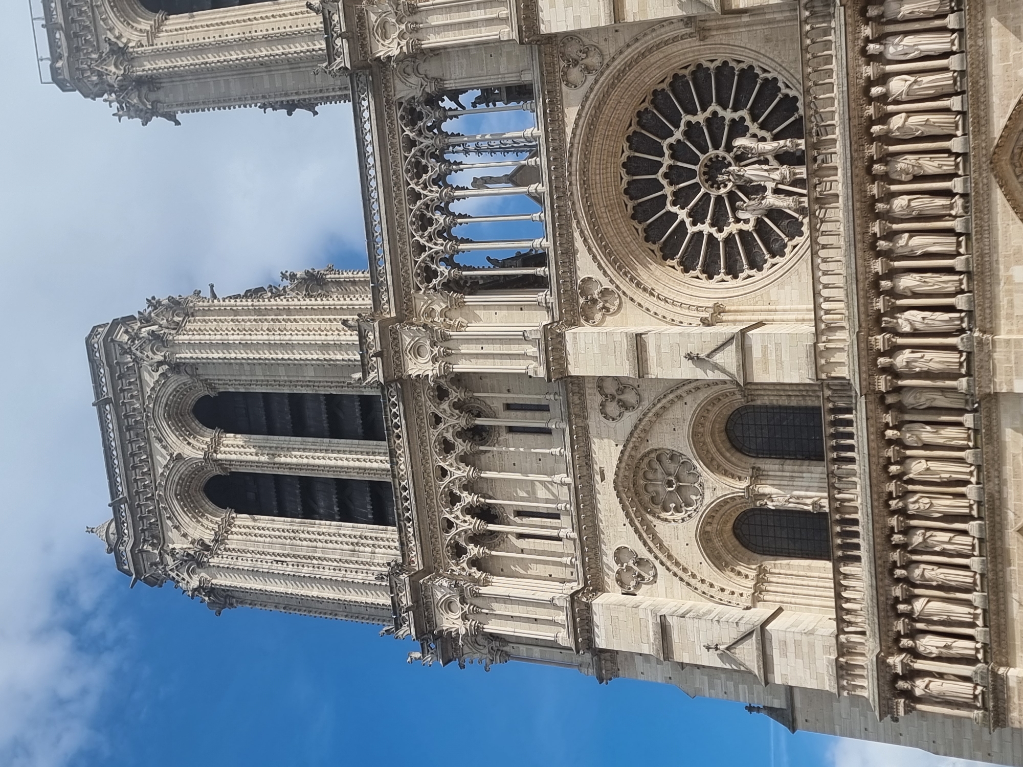 The image captures an upward-angled view of the magnificent Gothic facade of Notre-Dame Cathedral in Paris, France, against a bright blue sky with scattered white clouds. The perspective highlights the monumental scale and intricate architectural details of the historic structure.
The two western bell towers of the cathedral are visible, with the left tower prominent and a section of the right tower also shown. Connecting these towers is the Gallery of Chimeras, an open arcade adorned with numerous grotesque statues and finely carved balustrades. Below this gallery, a large, circular rose window dominates the central section, featuring elaborate stone tracery and a central grouping of figures.
To the right of the rose window, and extending horizontally across the facade, is the Gallery of Kings. This distinctive feature comprises a row of richly detailed statues, believed to depict the Kings of Judah, each set within its own arched niche. Throughout the facade, intricate carvings, decorative cornices, smaller arched windows, and additional gargoyles and grotesques can be seen. The stone appears in a light tan hue, illuminated by clear daylight. No specific activity is depicted, nor is any text visible in the image. The photograph effectively conveys the grandeur and historical artistry of this iconic Parisian landmark.