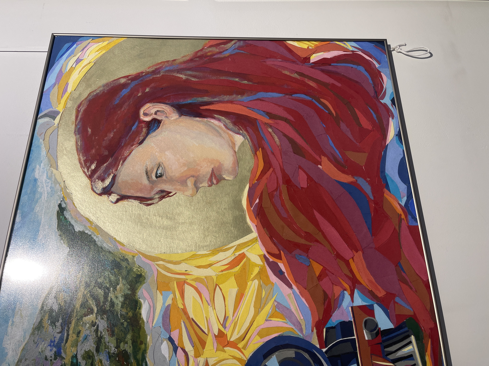 A framed artwork hangs on a white wall. The artwork depicts a person's face in profile, facing downward with red hair flowing around the head. Behind the head is a gold-colored halo. The artwork is composed of various colored pieces of paper or material, giving a mosaic-like appearance. The artwork seems to be indoors in a gallery or art space. There is no indication of Sofia, Bulgaria.