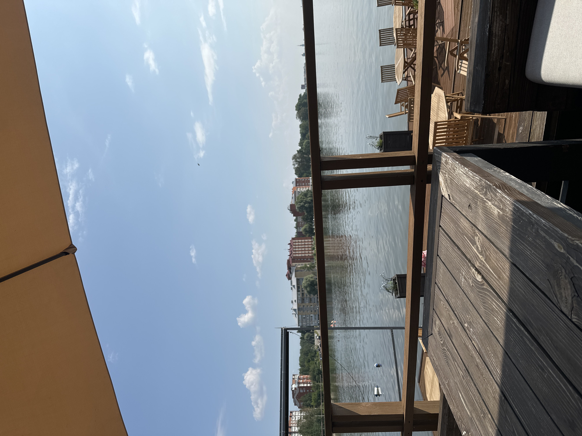 An outdoor seating area with wooden tables and chairs overlooks a body of water. In the foreground, a dark wooden table is prominent. Beyond it, several round and octagonal wooden tables are set with chairs on a wooden deck. The water is calm with some buoys visible. Across the water, a line of multi-story buildings, likely residential or commercial, is situated behind a treeline. The sky is blue with scattered white clouds, and the sun is shining, casting shadows on the deck. The scene appears to be a restaurant or cafe with a waterfront view in Votkinsk, Russia, possibly along the Votkinsk Reservoir.
