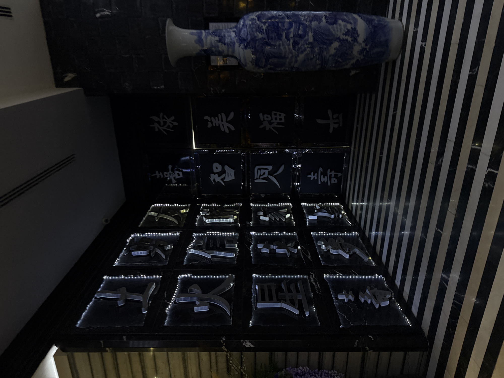 The image depicts an interior design element, possibly in a building in Sofia, Bulgaria. A large blue and white vase sits horizontally above a display of sixteen black square tiles, each featuring a stylized white character, possibly Chinese or Japanese, and outlined with small lights. Above these tiles are four larger tiles, each with a single white character. The display is situated near a wall with a black and white striped pattern. The overall color scheme is dark, with strong contrasts created by the white accents and lighting.