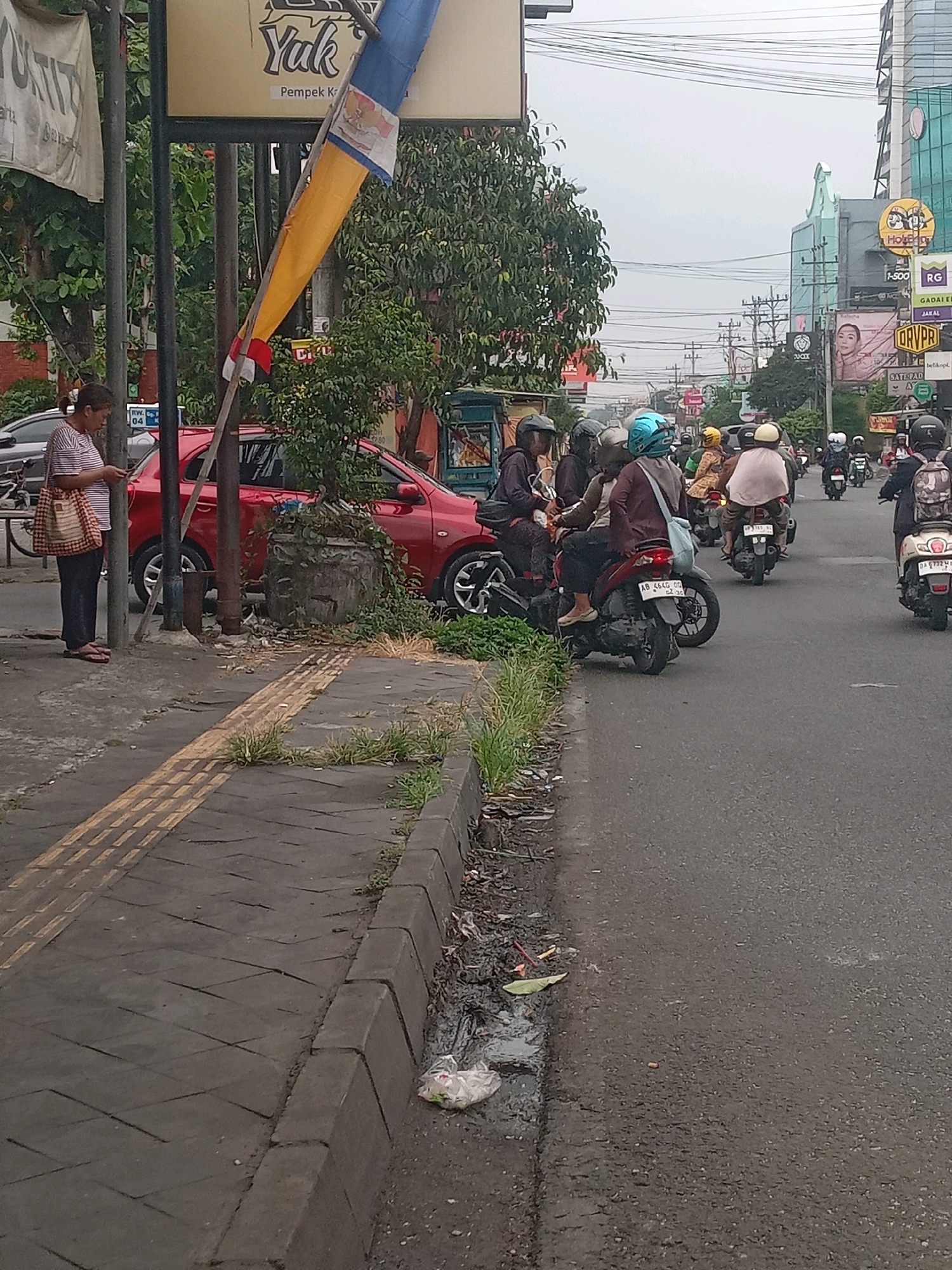 The image captures a street scene in Depok, Indonesia, on an overcast day. In the foreground, a sidewalk with tactile paving is visible, bordering a roadside drain filled with debris and some vegetation. A woman wearing a striped t-shirt and dark pants stands on the sidewalk, looking at her phone. Beside her, a red car is parked. A large banner hangs from a pole, displaying text that includes "Yuk" and "Pempek K".
Further down the street, a traffic scene unfolds with multiple motorcycles and riders. Several people are on motorcycles, some wearing helmets. The road is paved and shows signs of wear. Buildings and signs are visible in the background, indicating an urban environment. One building has a prominent green facade and a sign with "RG". Other signs include "1-500", "GARAL", and "ORVPRI". Overhead, a network of electrical wires crisscrosses the sky. The overall impression is a busy street in Indonesia, likely during daylight hours given the visibility.