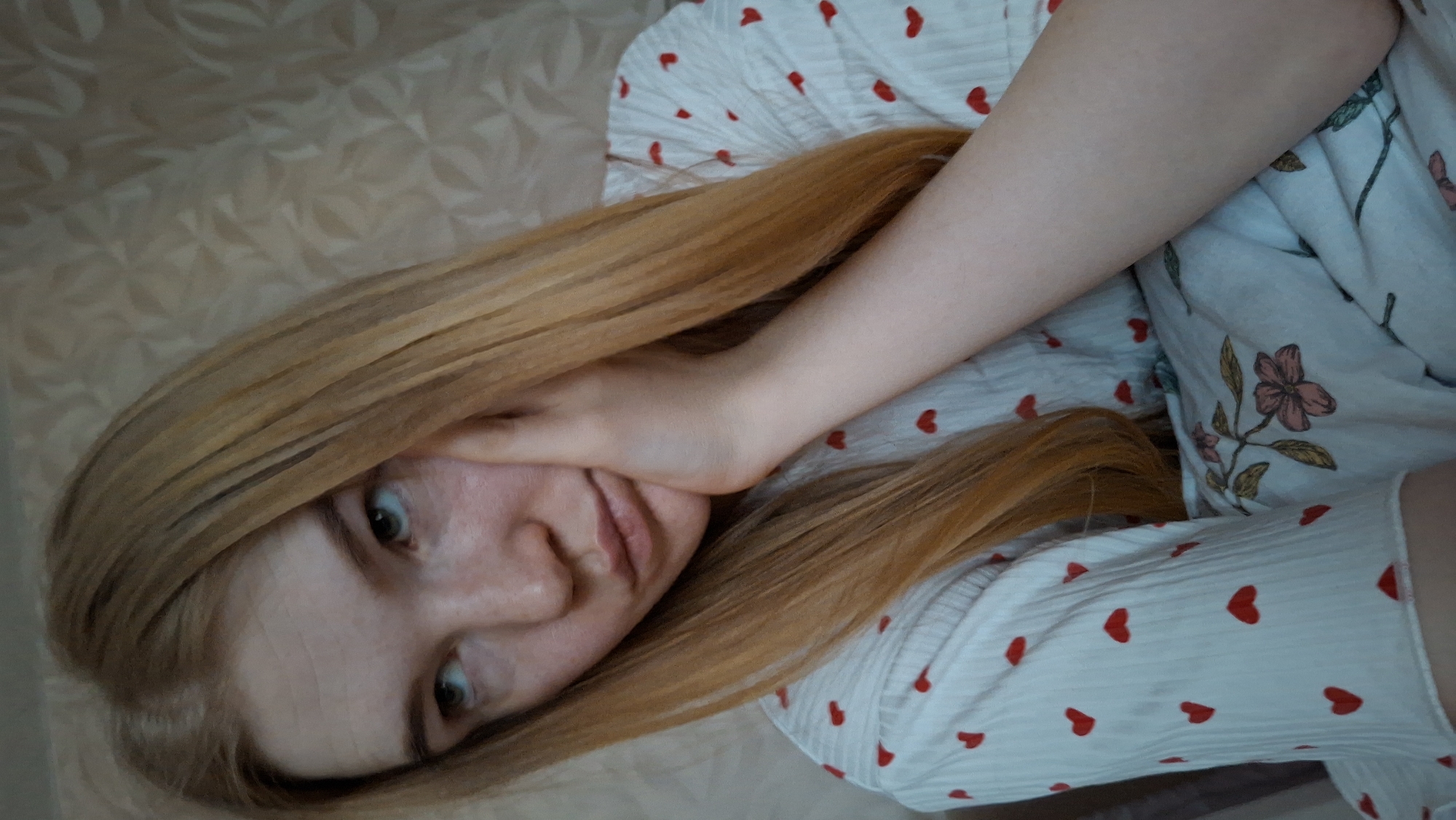 A woman with long, strawberry-blonde hair is lying down, possibly on a bed or sofa. Her right hand is positioned on her face. She is wearing a white pajama top patterned with small red hearts. A bedspread or blanket, also white, features red heart patterns and floral designs. The background shows a light beige or cream-colored surface with a subtle pattern. There is no definitive evidence to confirm the location as Chaikovsky, Russia.