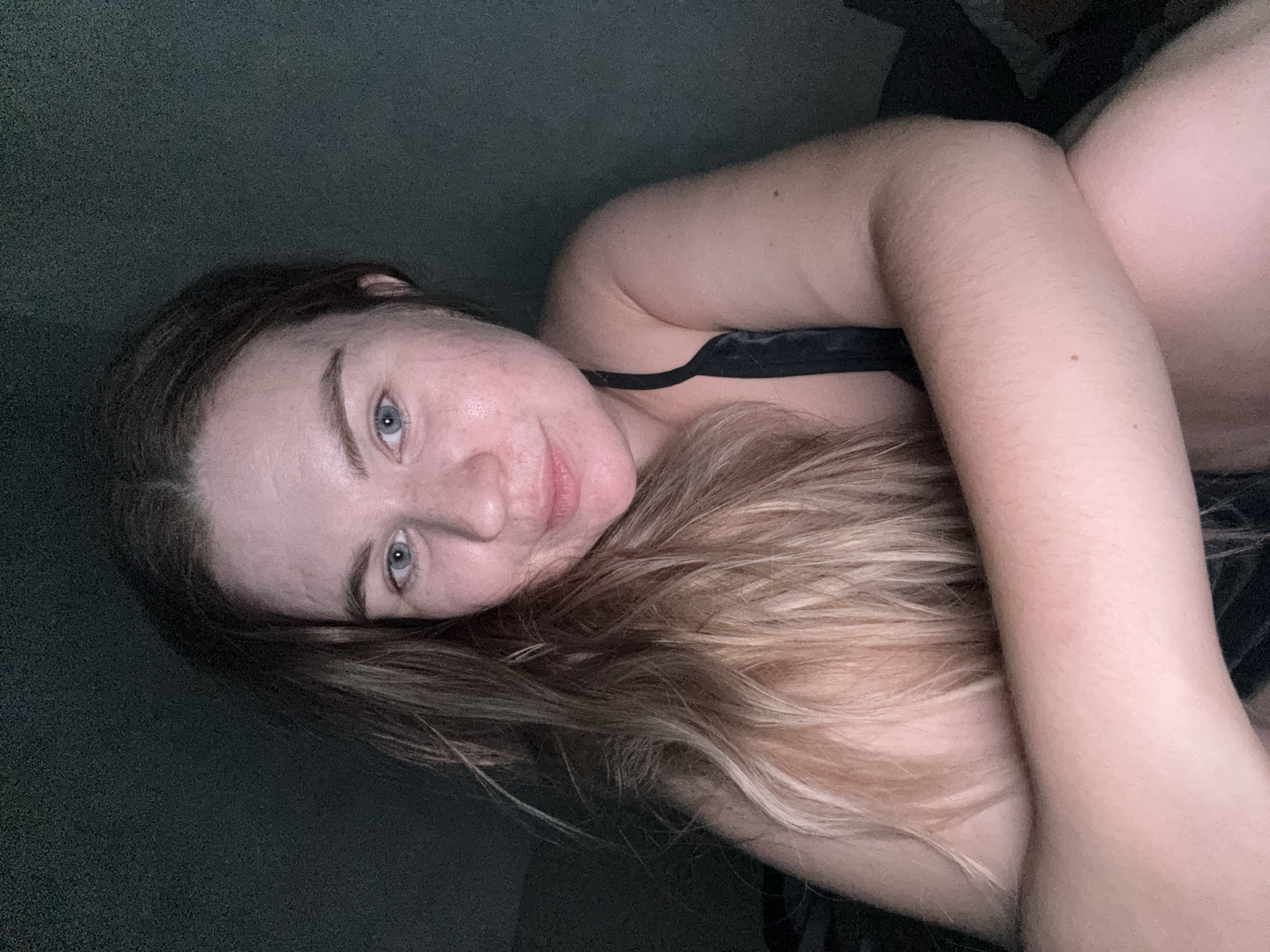 A woman with long, light brown and blonde hair lies on a dark surface, possibly a bed, in a dimly lit room. She has fair skin, blue eyes, and a small smile. She is wearing a black top or bra. The location is not discernible as Chaikovsky, Russia from this image. The image focuses on her face and upper body.