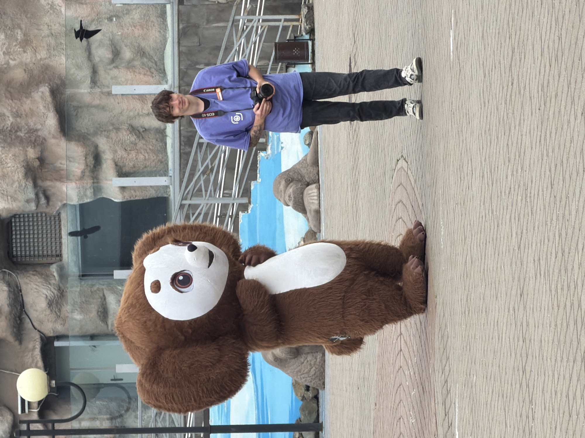 A person wearing a purple t-shirt, black pants, and white sneakers is standing and holding a camera. They have a Canon EOS 60 camera around their neck with the Canon logo visible. The person appears to be smiling. They are standing on a paved area.
In front of the person is a person in a mascot costume. The mascot is brown with a large white face, brown eyes, and a white oval on its chest. The mascot is also standing on the paved area. Behind the mascot and the person is a glass enclosure with a rock-like background and a blue sky with clouds painted on it. There are also some metal structures and a small dark object that appears to be a bird flying in the background. A yellow light fixture is visible in the lower left corner. The overall scene suggests an outdoor setting, possibly an zoo or amusement park. The lighting suggests it is daytime.