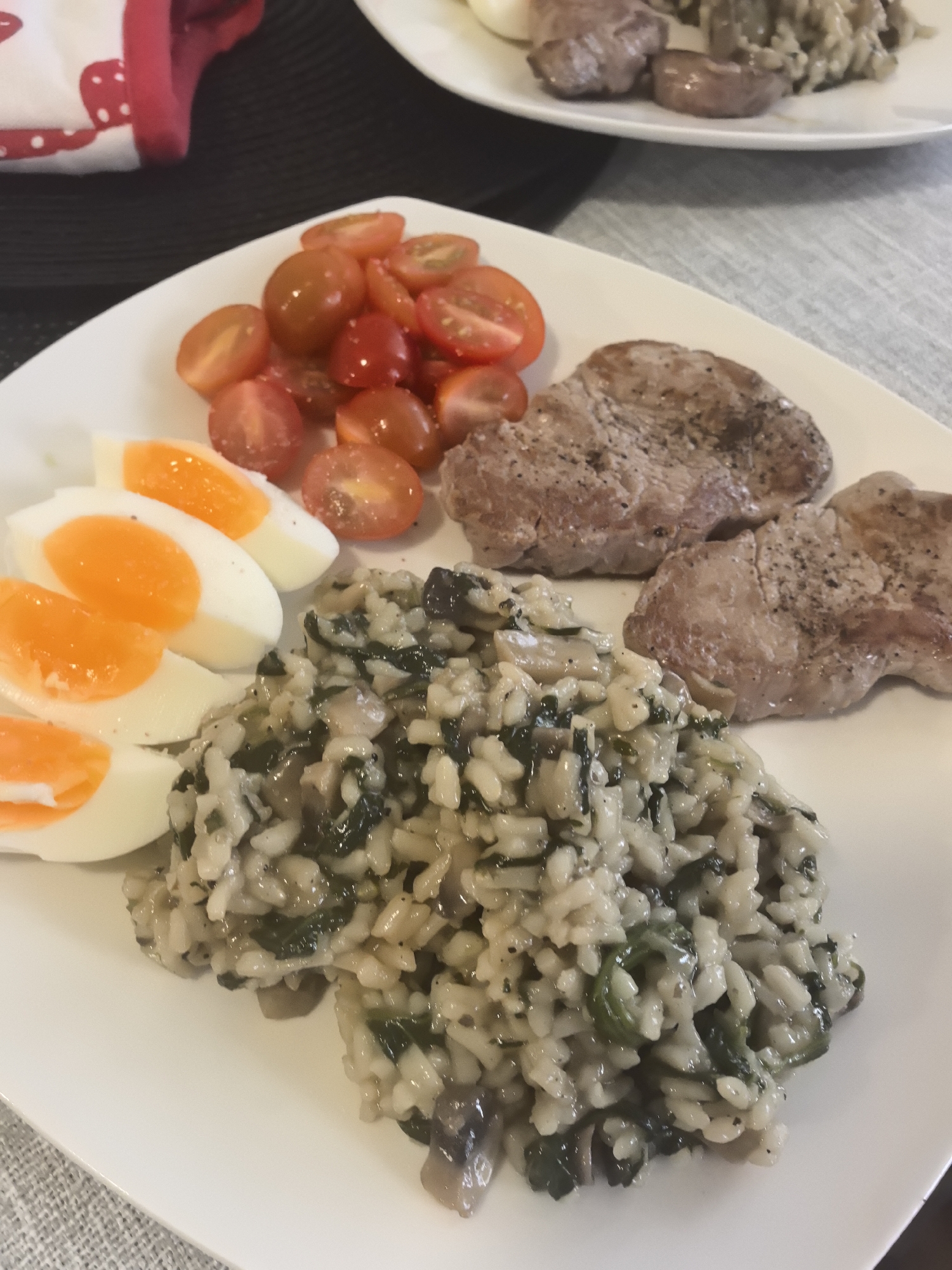 The image presents a plate of food, likely a meal, photographed in a domestic setting. The plate is white and contains the following items: three slices of hard-boiled egg with bright orange yolks, a pile of halved cherry tomatoes, two pieces of cooked meat, and a serving of spinach and mushroom risotto. Another plate with similar food arrangement is visible in the background. A red and white cloth is visible on the left side of the image. The plates are set on a grey table cloth. The precise location within Stara Zagora, Bulgaria is impossible to determine from the image alone.