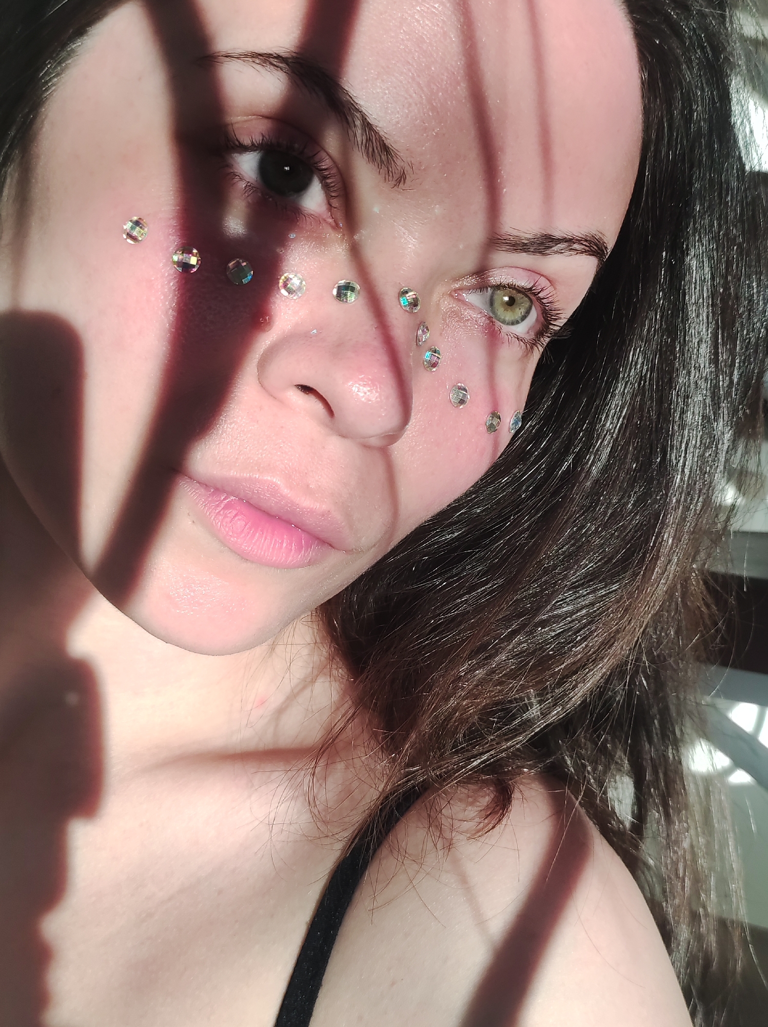A close-up portrait of a light-skinned young woman with light pink lips, dark hair and heterochromatic eyes (one brown, one green). She has small, iridescent adhesive gems decorating her face, primarily around her eyes and nose. The image is partially obscured by shadows, seemingly cast by a window frame or similar linear object. The woman is wearing a black top. The specific location in the city of Фоки, Russia, is not discernible from the image.