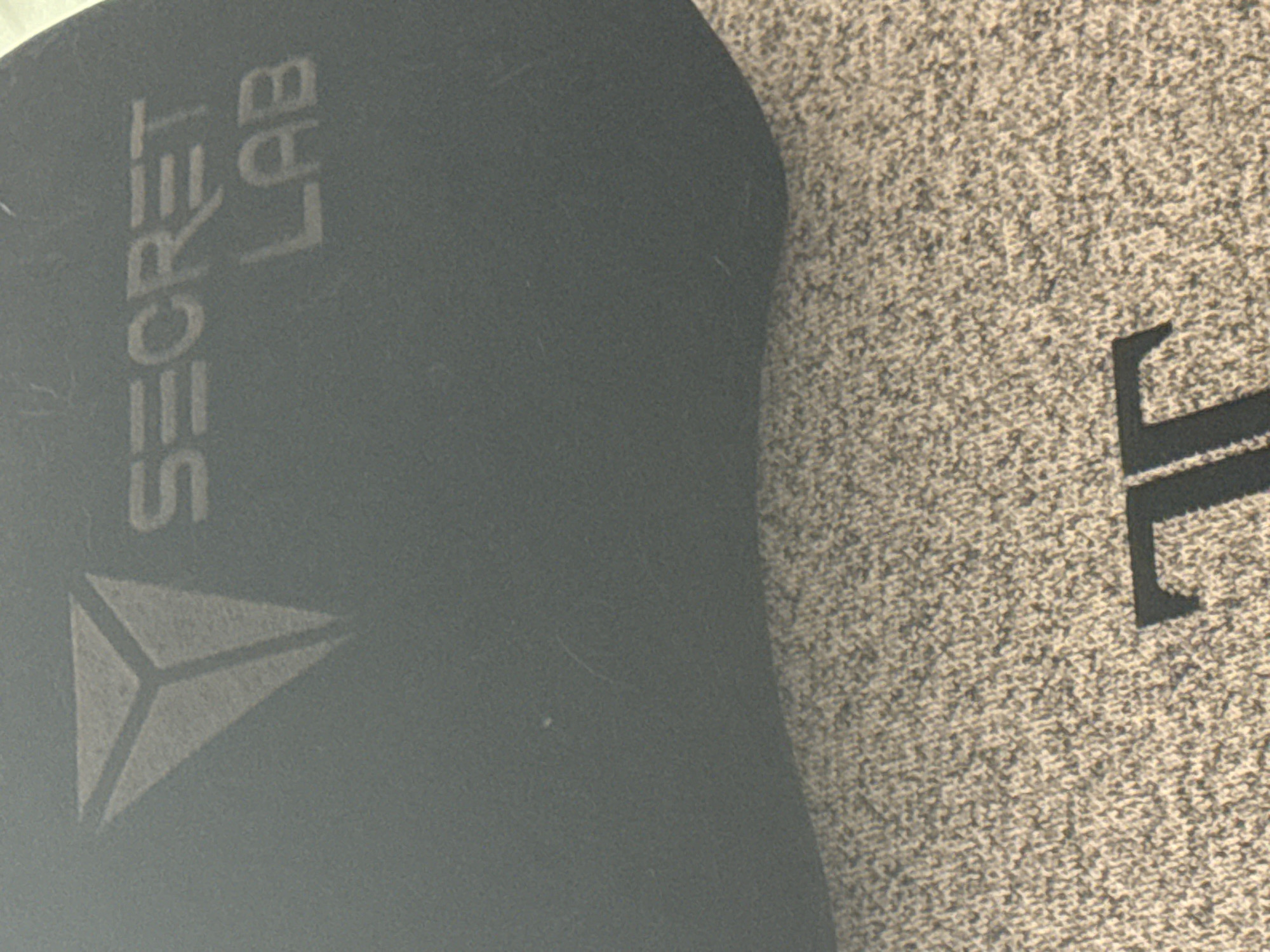 The image shows a close-up view of a dark gray fabric with a textured surface. On the fabric, a light gray logo in the shape of an inverted triangle with three lines inside is visible. To the right of the logo, the words "SECRET" and "LAB" are printed in the same light gray color. Below this, another textured surface with a dark gray "T" shape is visible. The image appears to be taken indoors, likely in a studio or retail environment. There are no people or animals visible in the image, and no specific location in Sofia, Bulgaria, can be identified from the visual information provided.