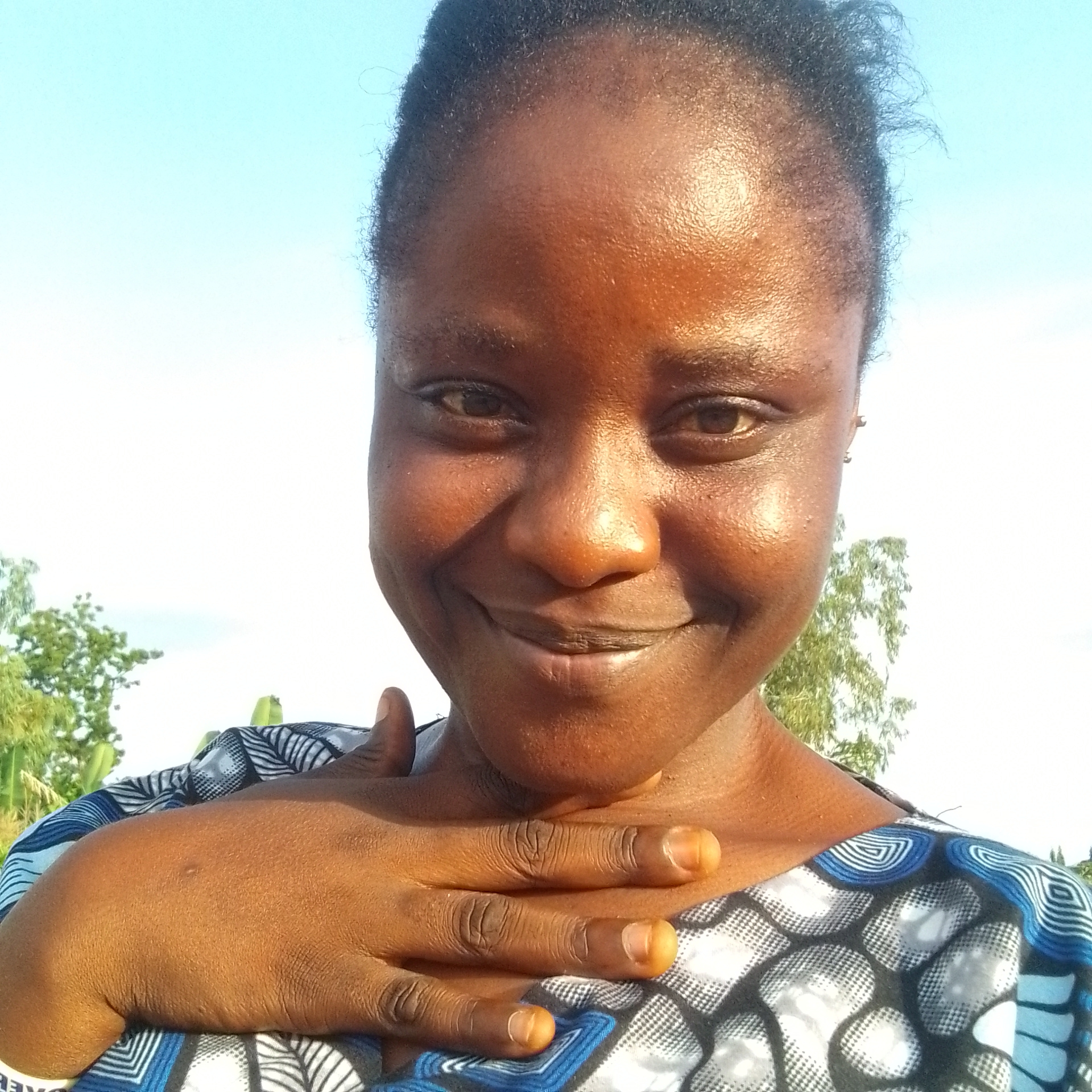 This is a close-up selfie of a Black woman, taken outdoors in Jalingo, Nigeria. She is looking directly at the camera with a gentle smile, and her left hand is resting on her chest. Her skin is a warm brown, and her hair is dark and pulled back. She is wearing a patterned top with blue, white, and black designs. The background is a clear blue sky, with some greenery and trees visible on the sides, suggesting it is daytime and likely sunny. The overall impression is one of warmth and contentment. There is no visible text in the image.