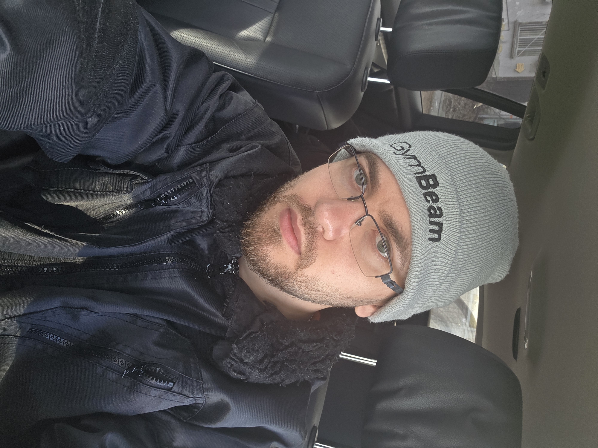 A man with fair skin, facial hair, and glasses wearing a grey "GymBeam" beanie and a black jacket with a fur collar, takes a selfie inside a vehicle. The interior of the vehicle includes black leather seats. The background shows a blurred view of what appears to be a building, possibly indicating an urban setting. The location is unknown, as I cannot identify the city of Plovdiv, Bulgaria, based on the image alone.