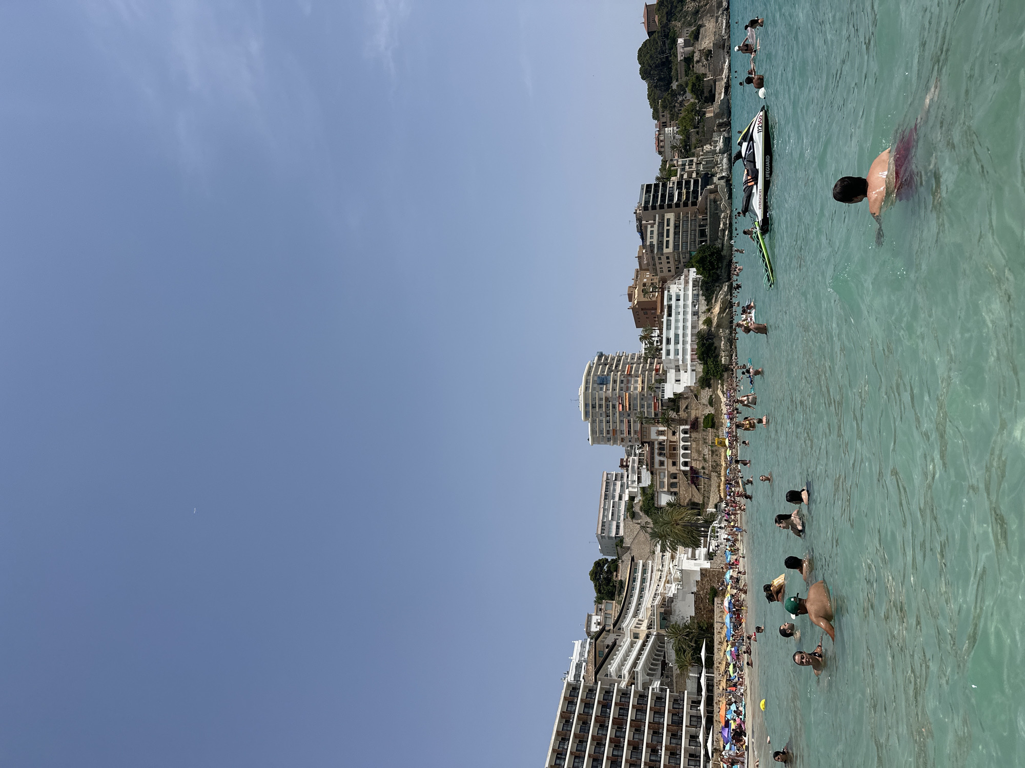The image depicts a crowded beach scene in Sant Agustí, Spain, under a clear blue sky with a faint airplane contrail visible. The turquoise water is filled with numerous people swimming and wading. In the foreground, a man in red swimming trunks swims away from the camera. Further out, a jet ski with "EMERGENCY SERVICES" and "Lifeguard" written on it is docked with its green tube. The beach itself is densely populated with sunbathers relaxing under colorful umbrellas and families enjoying the water. Behind the beach, a hillside is densely packed with multi-story white buildings, indicative of a tourist resort, with balconies and windows facing the sea. Palm trees are interspersed among the buildings, and a few more buildings are visible on the higher parts of the hills. The overall scene conveys a lively and popular summer vacation destination.