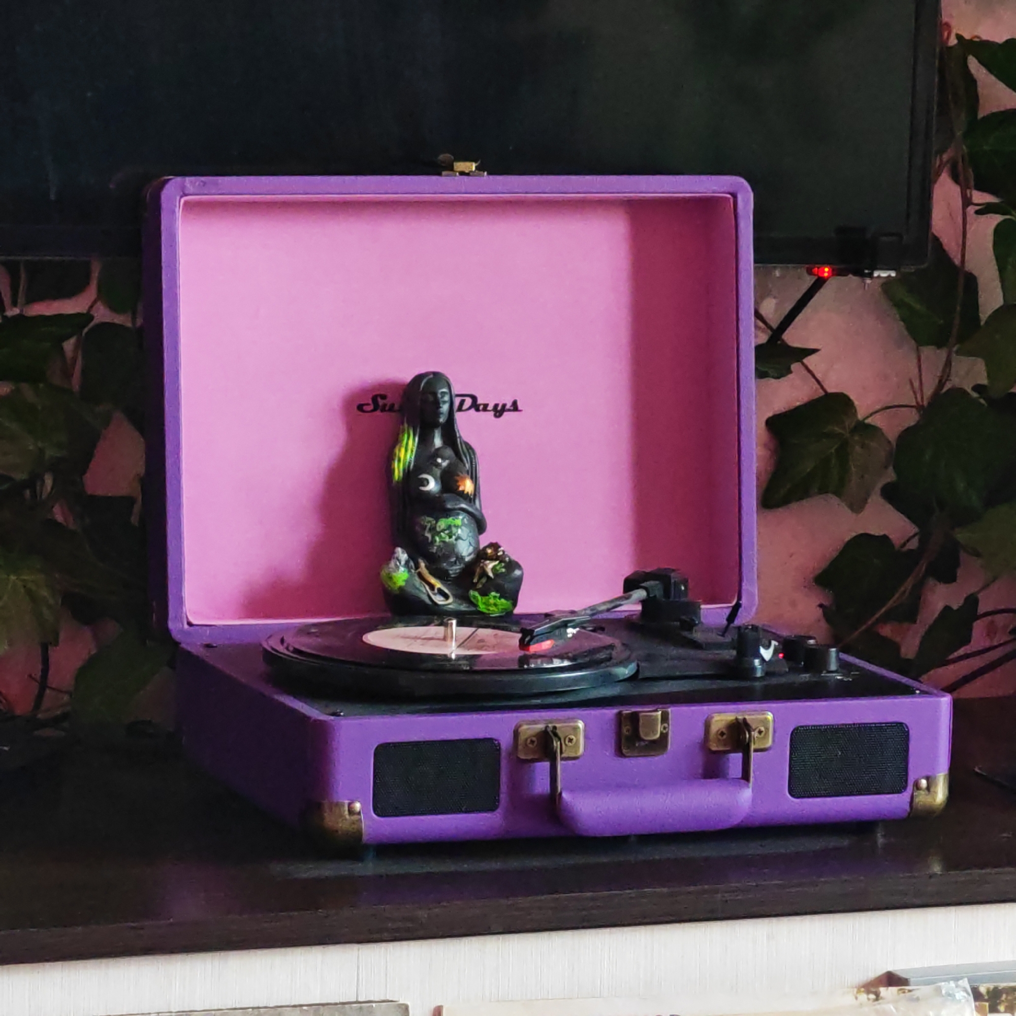 A slightly high-angle, medium shot depicts an open, purple and pink suitcase-style record player on a dark surface, likely a table or shelf. A black vinyl record is on the turntable, and a dark statue of a figure holding an object is standing near the center of the vinyl. The open lid of the record player reveals a pink interior with the text "Sun Days" printed on it. Behind the record player, a monitor or TV screen is partially visible on the left, and lush green foliage is on the right. The location is identified as Rostov, Russia, according to information not visible in the image.
