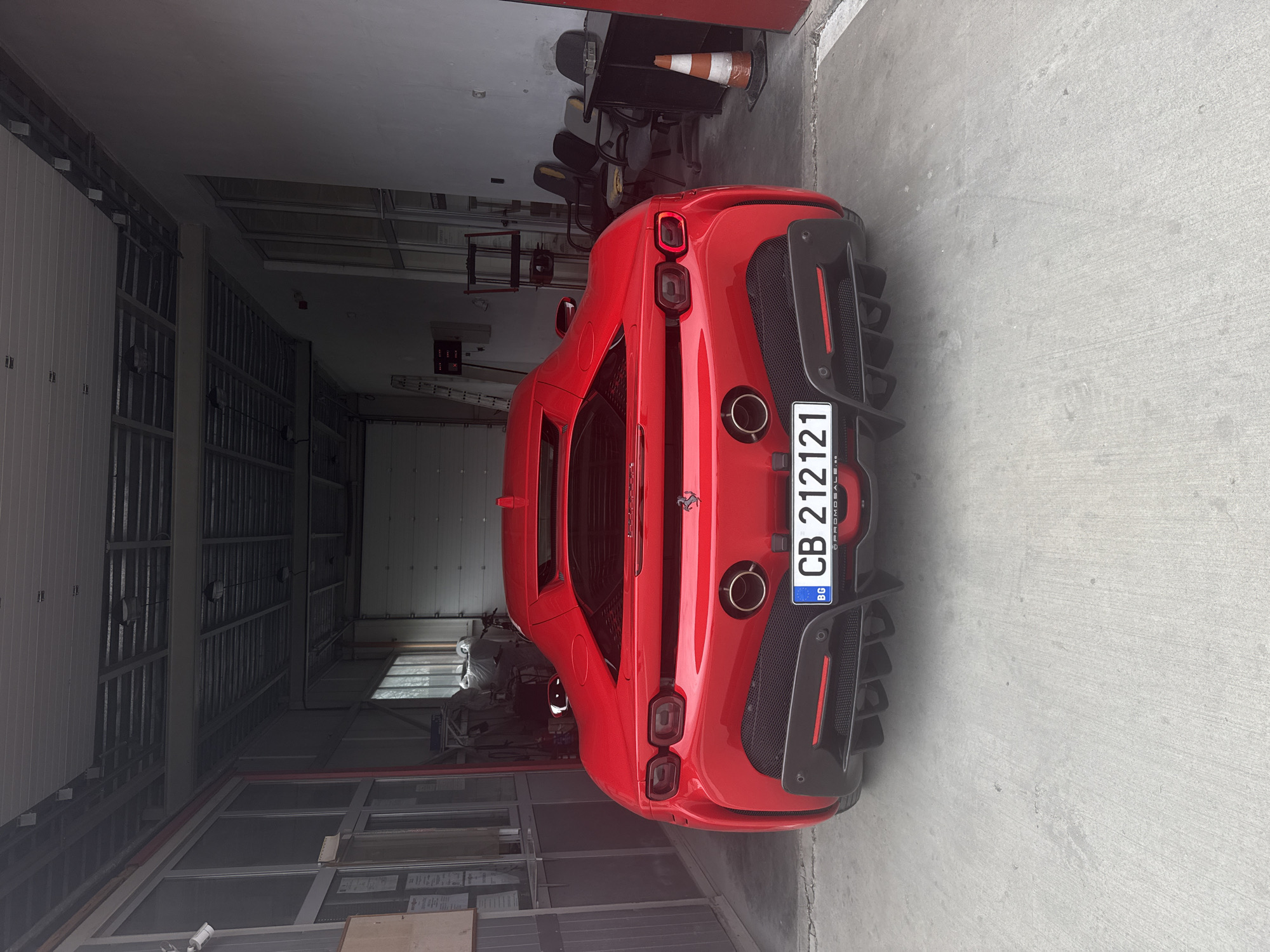 A red Ferrari is parked in a garage. The car has a Bulgarian license plate that reads "CB 212121." The garage appears to be located in an industrial or commercial area, possibly in Plovdiv, Bulgaria, given the license plate. Other items like chairs and storage are visible in the garage.