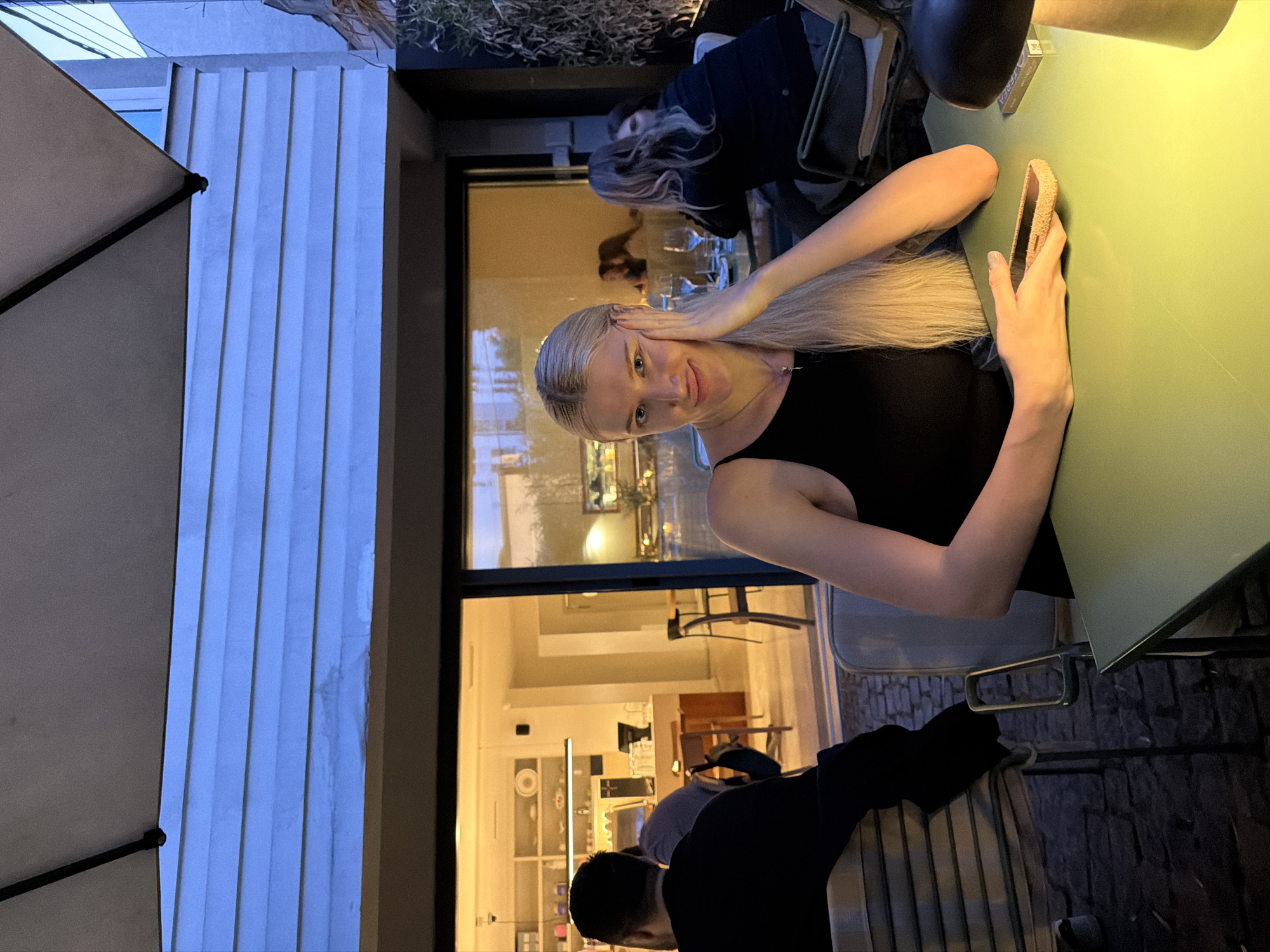 The image shows a blonde woman seated at a table outdoors, possibly at a restaurant in Belgrade, Serbia. She is wearing a black sleeveless top and is holding a cell phone in her right hand, with her left hand near her head. The table is green, and the lighting suggests it is evening. Other people are visible in the background, within the restaurant's interior and at another table. There is a white awning to the left, and the restaurant has large glass windows. The interior of the restaurant is well-lit and shows what appears to be a bar and dining area.