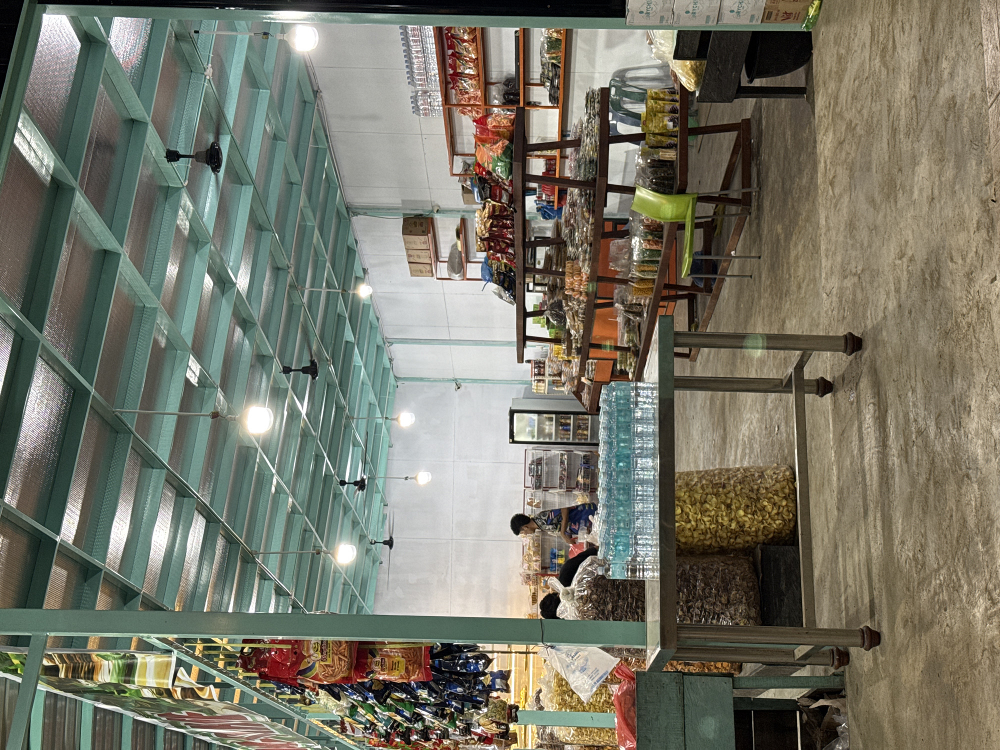 The image depicts the interior of a store located in Grong-Grong, Indonesia. The store appears to be a small, local market or convenience store, with shelves stocked with various food items, snacks, and beverages. In the foreground, a metal table holds rows of bottled water. To the left, large bags of what appear to be snacks are visible. In the background, a person wearing a blue and white patterned shirt is standing near shelves filled with packaged goods. Another person is partially visible to the left of the main figure, facing away from the camera. The store features a teal-colored metal frame supporting the roof, which is covered with insulation panels. Ceiling fans are mounted on the roof. The lighting is provided by overhead bulbs. The floor is made of concrete. The store's exterior is not visible, but the interior suggests it is a functional retail space.