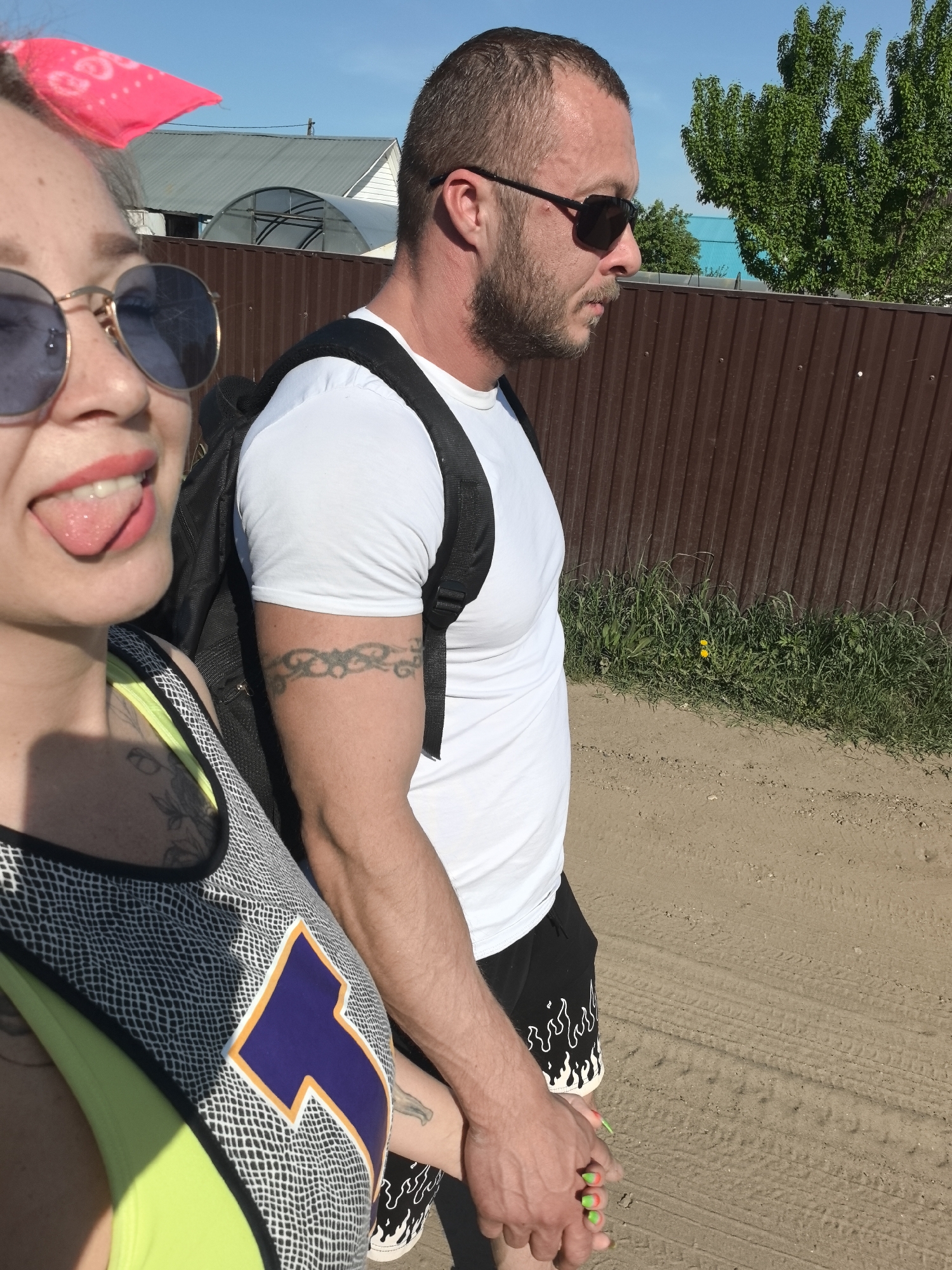 A couple is walking on a dirt road in Noviy, Russia, holding hands. The woman is on the left, wearing round sunglasses, a pink headscarf, and a neon green and patterned top. She has her tongue sticking out. The man on the right is wearing sunglasses, a white t-shirt, and black shorts with a flame pattern. He has a black backpack and a tattoo on his left arm. They are walking next to a brown corrugated metal fence. In the background, there are trees and buildings. The ground is sandy with tire tracks.