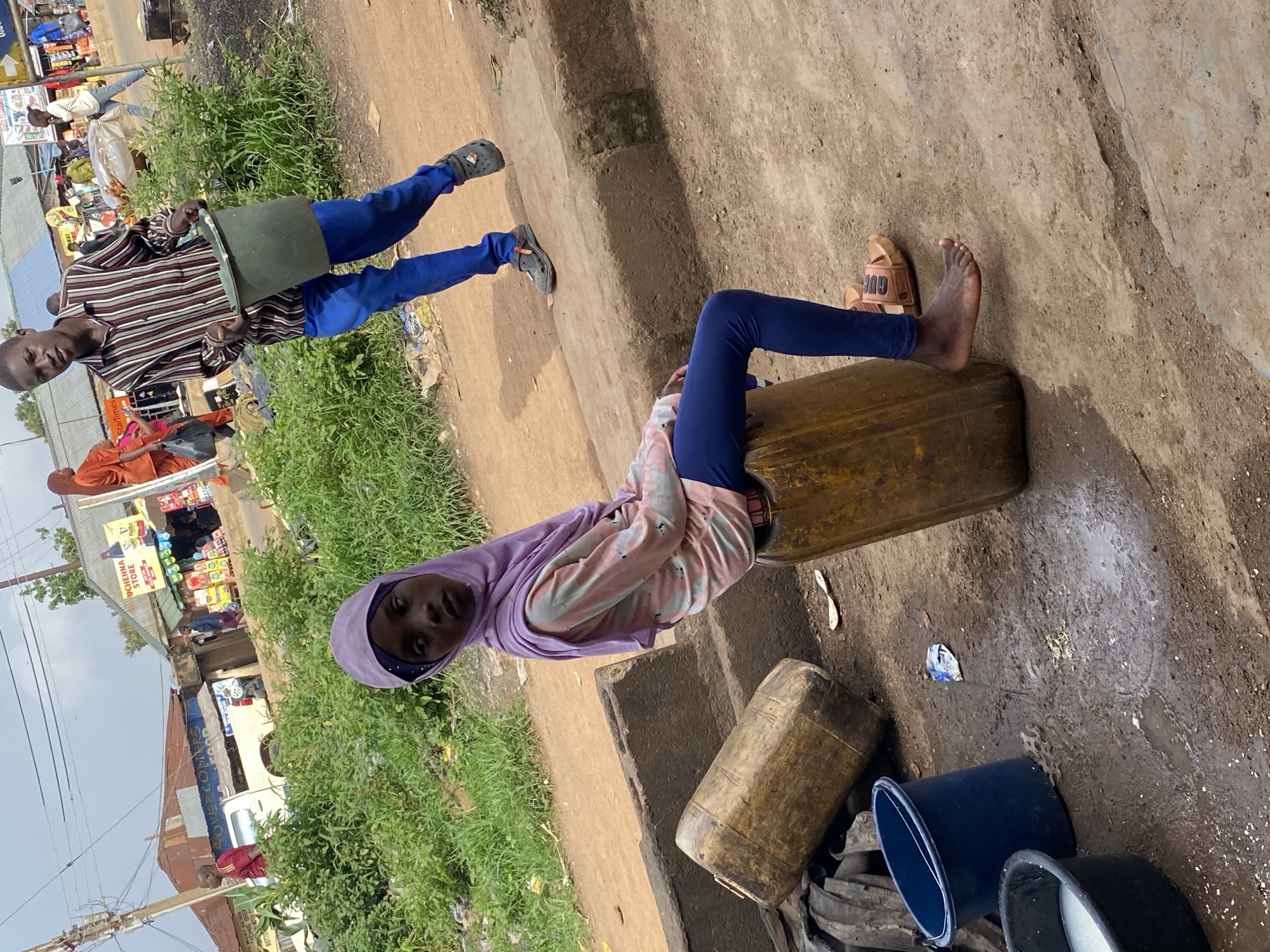 In Bukuru, Nigeria, a young girl wearing a lavender hijab and a multi-colored long-sleeved shirt sits on a large yellow plastic barrel on a dirt road. She has dark skin and is wearing blue leggings and brown sandals. Beside her, a large wooden barrel lies on its side. In the foreground, a blue plastic bucket and a black basin containing a white liquid are visible. In the background, a young man in a striped shirt and blue pants walks away from the camera, carrying a green bucket. Other people and market stalls are visible further in the background. The ground is dusty and appears wet in some areas, with scattered debris.