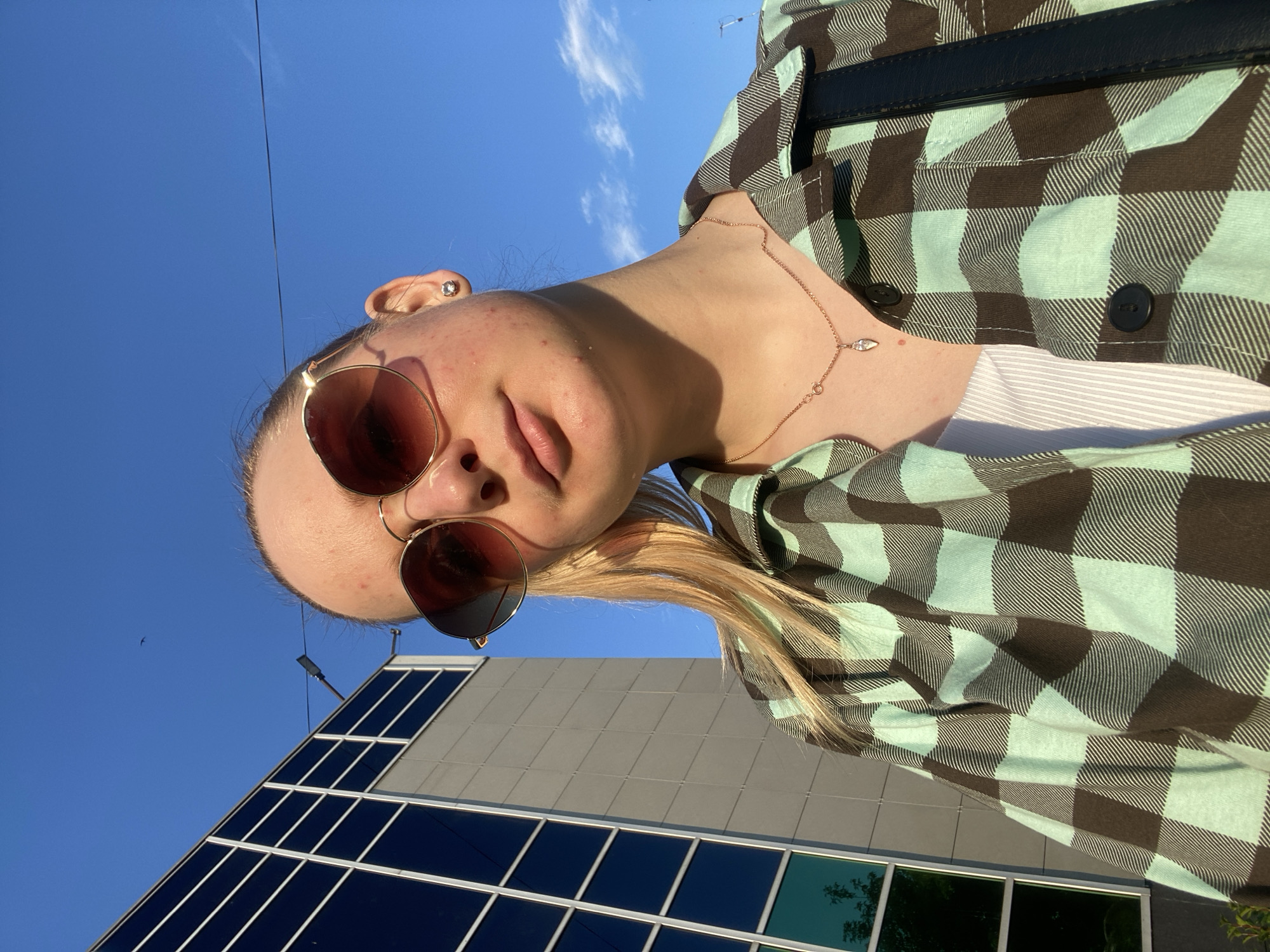 A young woman stands outdoors, taking a selfie. She has blonde hair pulled back in a ponytail and is wearing large, round sunglasses with a rose-gold tint. Her face is turned slightly towards the camera, and she is looking at it. She has on a light blue and brown plaid shirt over a white top, and a delicate rose-gold necklace with a diamond pendant. She is also wearing a small stud earring in her visible ear. Behind her, a modern building with a glass facade and blue sky are visible. The lighting suggests it is daytime, likely late afternoon, with the sun casting shadows and highlighting parts of her face and clothing.
