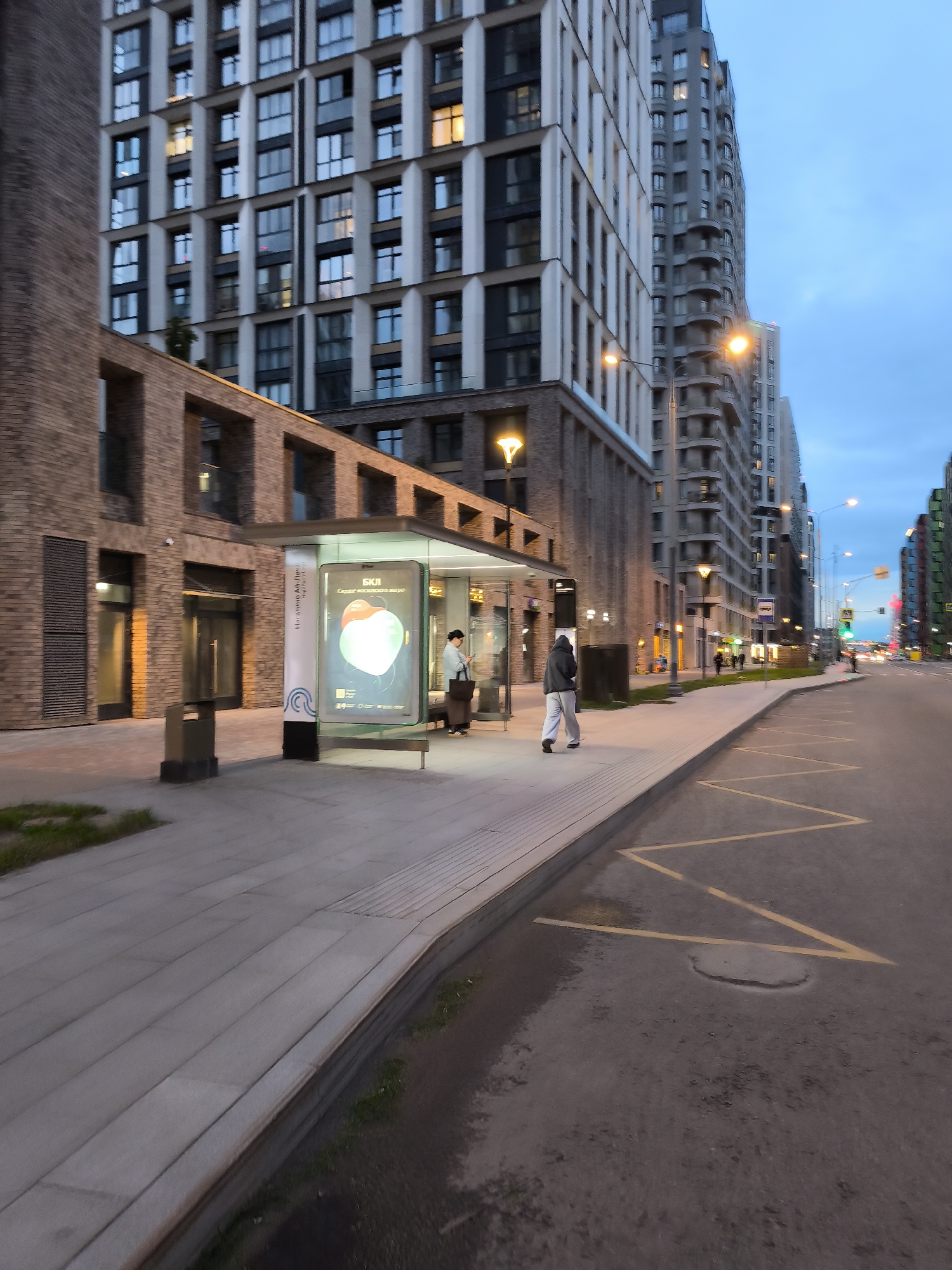 The image depicts a street scene in Moscow, Russia, at dusk. A modern bus stop is situated on the sidewalk, featuring illuminated advertisements. Two individuals are present: one is seated inside the bus stop, and another is walking away from it along the sidewalk. The surrounding architecture consists of tall, modern buildings with a mix of brick and concrete facades. Streetlights illuminate the scene. The road is visible with yellow parking restriction markings. The sky is a blend of blue and gray, indicating the time of day. The overall atmosphere suggests a quiet urban environment.