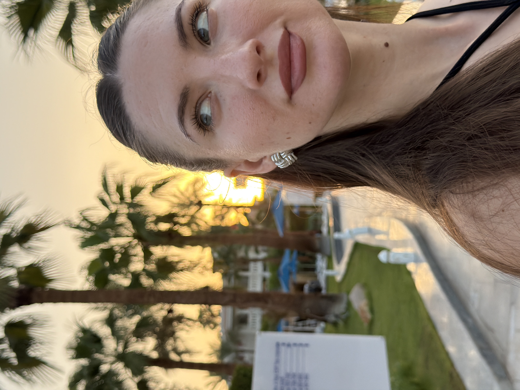 A woman with long brown hair and light skin is posing for a photo outdoors at sunset in Sharm El Sheikh, Egypt. She is wearing a black top and silver earrings. In the background, there are palm trees, a white building, and a pathway. The sun is setting behind the palm trees, casting a warm glow over the scene.