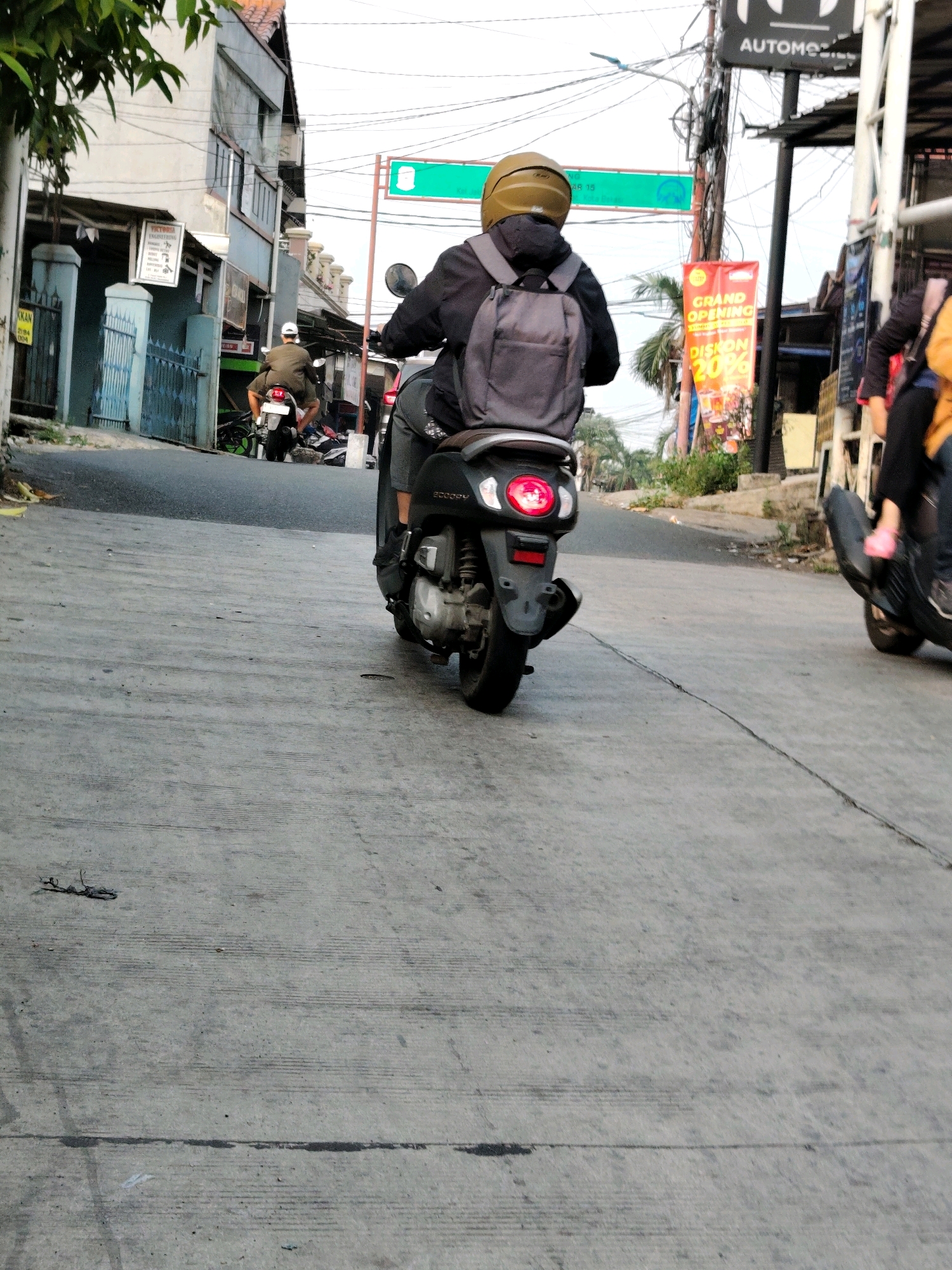 The image depicts a street scene in Bekasi, Indonesia, with a focus on several motorcycles and their riders. In the foreground, a black motorcycle is parked on the right side of the road, viewed from the rear. A person wearing a dark jacket and a yellow helmet is riding a black motorcycle in the middle of the road, also seen from behind. This rider is carrying a gray backpack. Further down the street, another person on a motorcycle is visible, wearing a light-colored cap and a dark outfit. Various signs are present along the street, including a green traffic sign and a red banner advertising a "GRAND OPENING" with "DISKON 20%." Buildings line the street, with visible architectural details such as windows and a tiled roof on the left side. The road surface is asphalt with visible cracks and markings. Trees and foliage are also present, contributing to the urban environment. The sky is overcast.