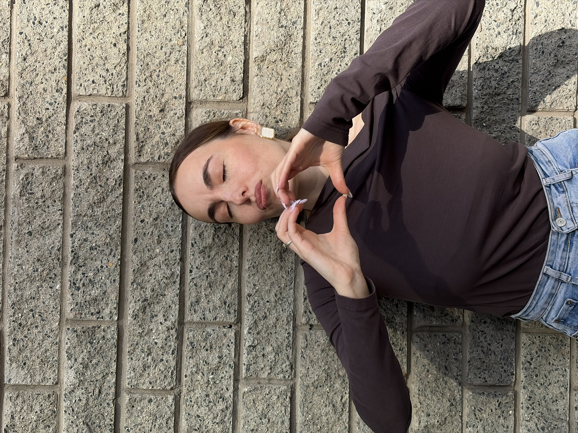 The image shows a woman lying on a concrete wall in an outdoor setting. She is wearing a dark brown long-sleeved shirt and light blue jeans. Her eyes are closed, and she is holding her hands in a heart shape near her face. She wears square earrings. The wall is made of rectangular concrete blocks. Based solely on the image, the location in Izhevsk, Russia cannot be determined.