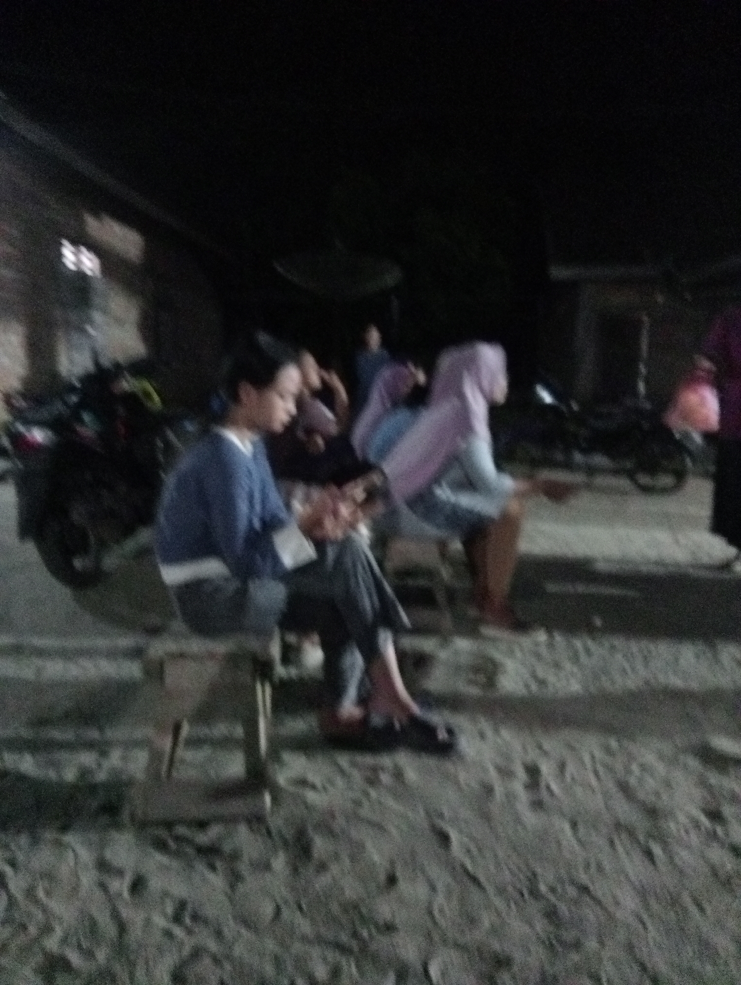 The image depicts a dimly lit outdoor scene, possibly a roadside or gathering area in Rasuan Baru, Indonesia. In the foreground, a young person wearing a blue long-sleeved shirt and dark pants sits on a wooden bench, looking down at something in their lap. To their right, another individual wearing a light purple hijab and a light blue garment is also seated, facing away from the viewer. Behind them, motorcycles are parked, and other individuals are visible in the background, though their features are obscured by the low light and blurriness of the image. The ground appears to be uneven and possibly sandy or dusty. The overall impression is of a casual gathering or waiting area in a somewhat informal setting.