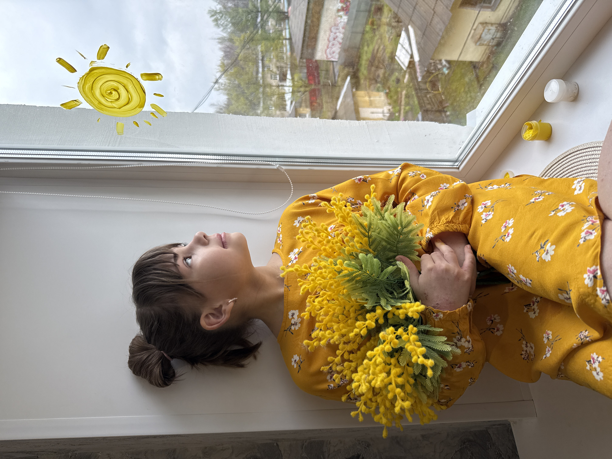 A young woman is lying on a windowsill, gazing upwards, with her head near a window. The woman has fair skin, brown hair styled in a low bun, and wears a yellow dress with a white floral pattern. She is holding a bunch of yellow flowers. The window has a painted yellow sun on the glass. Outside, there is a view of buildings and trees, indicative of an urban environment. The location is Rusanovo, Russia. There are jars on the right side of the windowsill.