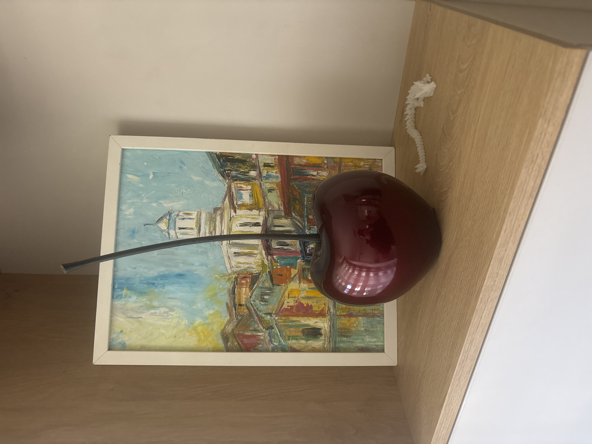 The image presents a close-up, slightly angled view of a wall or shelf arrangement. The foreground contains a framed painting of what appears to be an abstract cityscape. Dominating the scene is a large, glossy, burgundy-colored sculpture of a cherry with a black stem. Adjacent to the painting is a light brown wooden surface, likely a cabinet or shelf, and further up is a light cream wall. A decorative white paper string is mounted on the wooden surface. The location cannot be determined to be Plovdiv, Bulgaria with the given information.