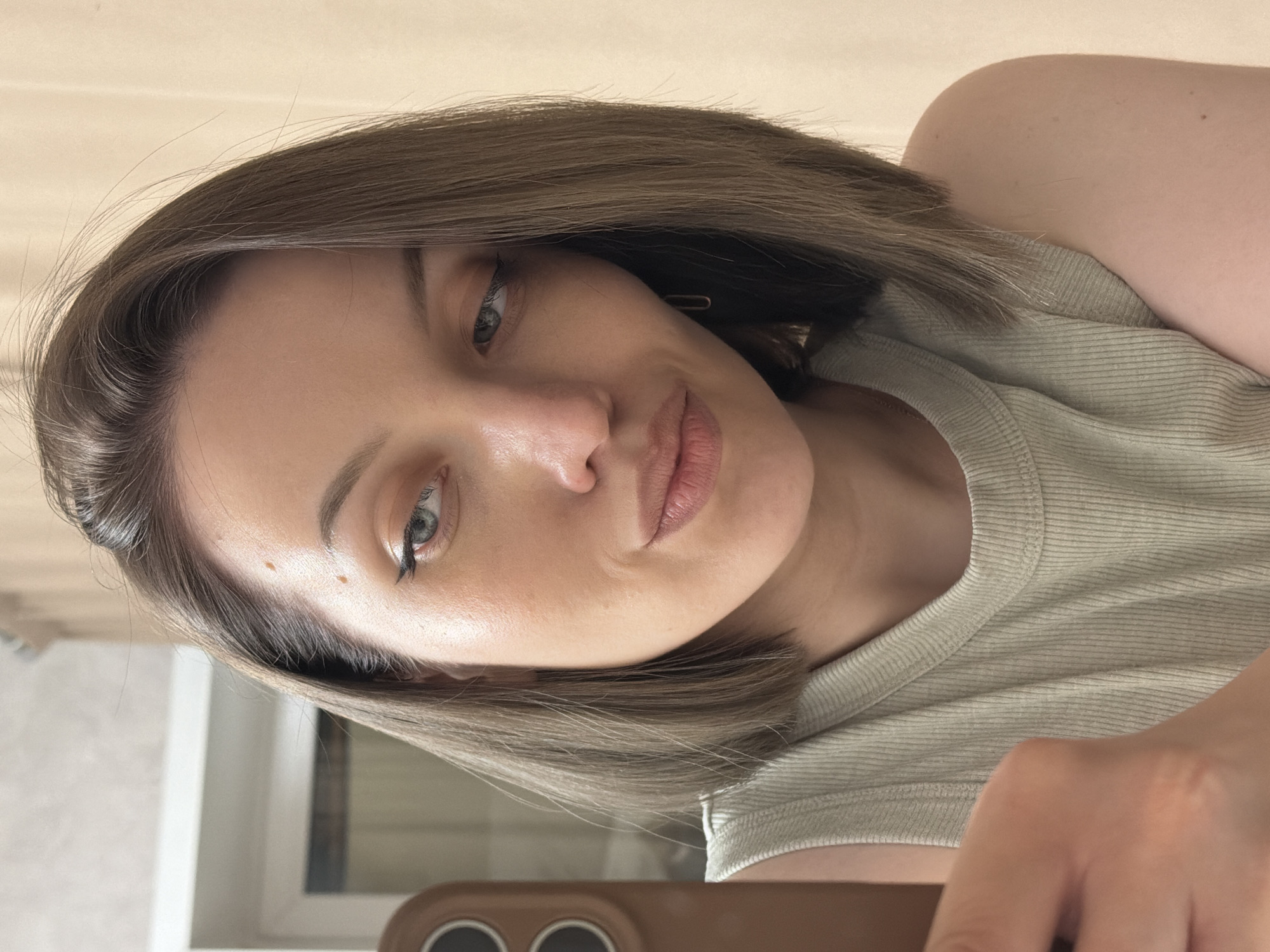 A young woman is shown in a close-up, angled selfie. She has fair skin, gray eyes, and short, light brown hair. She is wearing a light green ribbed tank top. She is holding a brown phone in her hand, which is visible in the lower portion of the image. A window is visible in the background. The location is not discernible as being in Reshetnikovo, Russia, based on the visual information in the image.