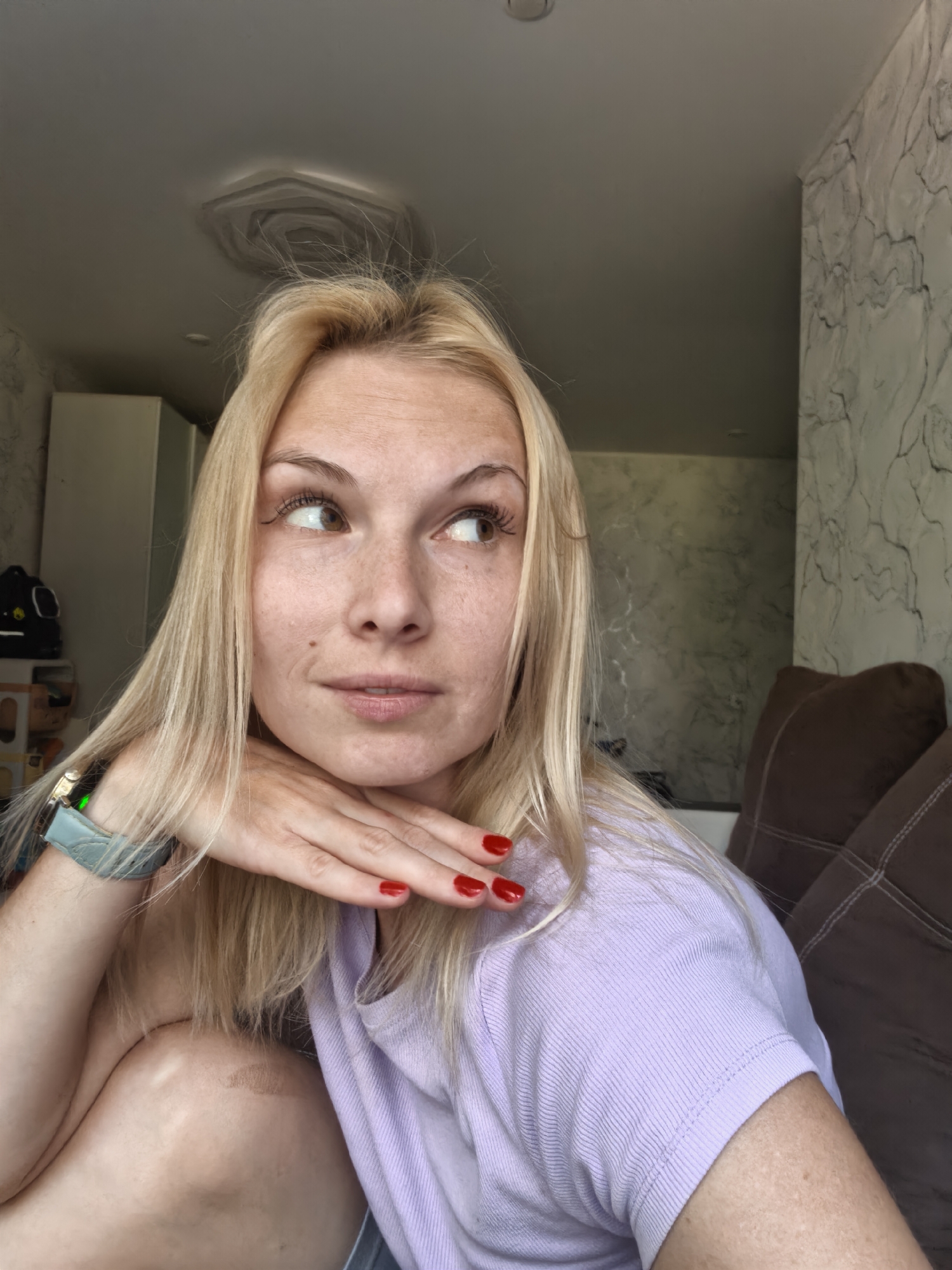 A blonde woman is resting her chin on her hand, looking to the right. She has blonde hair, brown eyes, and freckles. She is wearing a light purple t-shirt and a watch with a blue strap. Her fingernails are painted red. The background appears to be an indoor setting, possibly a living room, with a couch and a marble-patterned wall. There is a ceiling with an unusual textured design. The scene does not depict any elements that would allow for localization to the city of Sterlitamak, Russia.