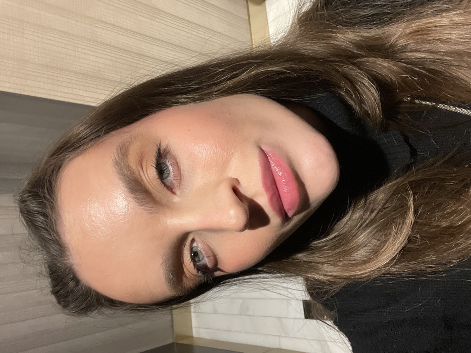 A woman with light skin, long brown hair, and blue eyes is pictured close-up. Her face is partially illuminated, highlighting her smooth complexion and glossy pink lips. She wears a black turtleneck sweater. The background includes what appears to be a light-colored wooden surface and a white brick wall. The image is taken from a tilted perspective, with the woman's face angled towards the viewer.