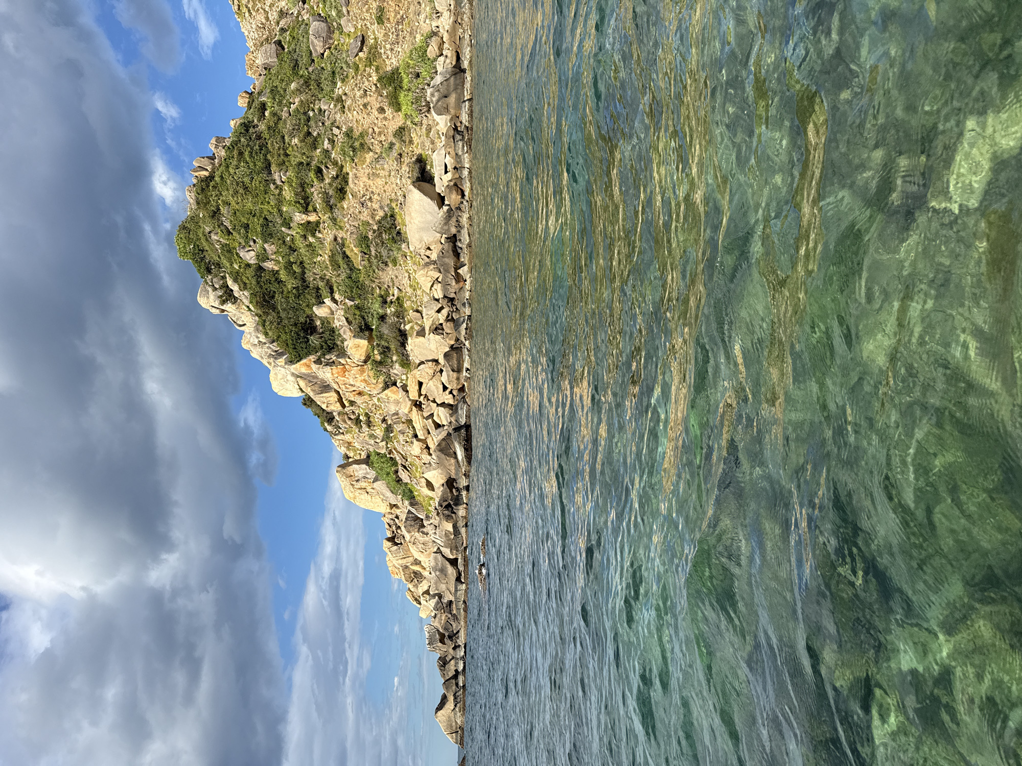 The image captures a coastal scene with a rocky island covered in sparse vegetation in the background. The foreground is dominated by clear, green water with visible ripples and reflections. The sky above is partly cloudy, with patches of blue visible. The rocky island features large boulders and a mix of brown earth and green plant life. The water appears shallow near the shore, allowing the seabed to be seen in places. The overall impression is one of a natural, undeveloped coastal environment. The location is identified as Từ Nham, Vietnam.