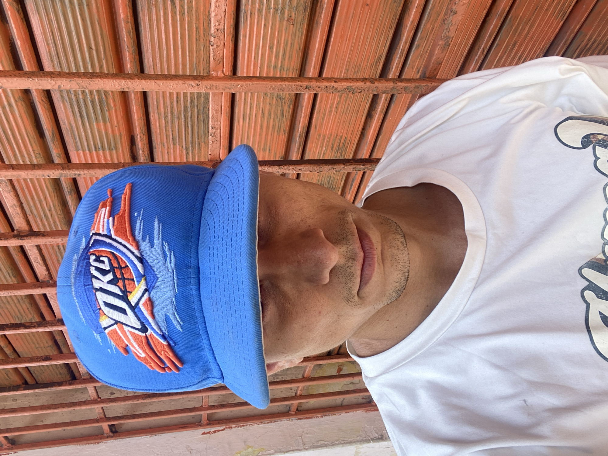 A medium-shot shows a man looking down at the camera with a blue baseball cap that has the logo "OKC" on the front, wearing a white t-shirt, positioned under a corrugated orange metal ceiling. The location appears to be in an outdoor or semi-outdoor space, indicated by the structure above him. While the general environment is visible, there is no explicit information within the image itself to confirm the location is specifically San José de Barlovento, Venezuela.
