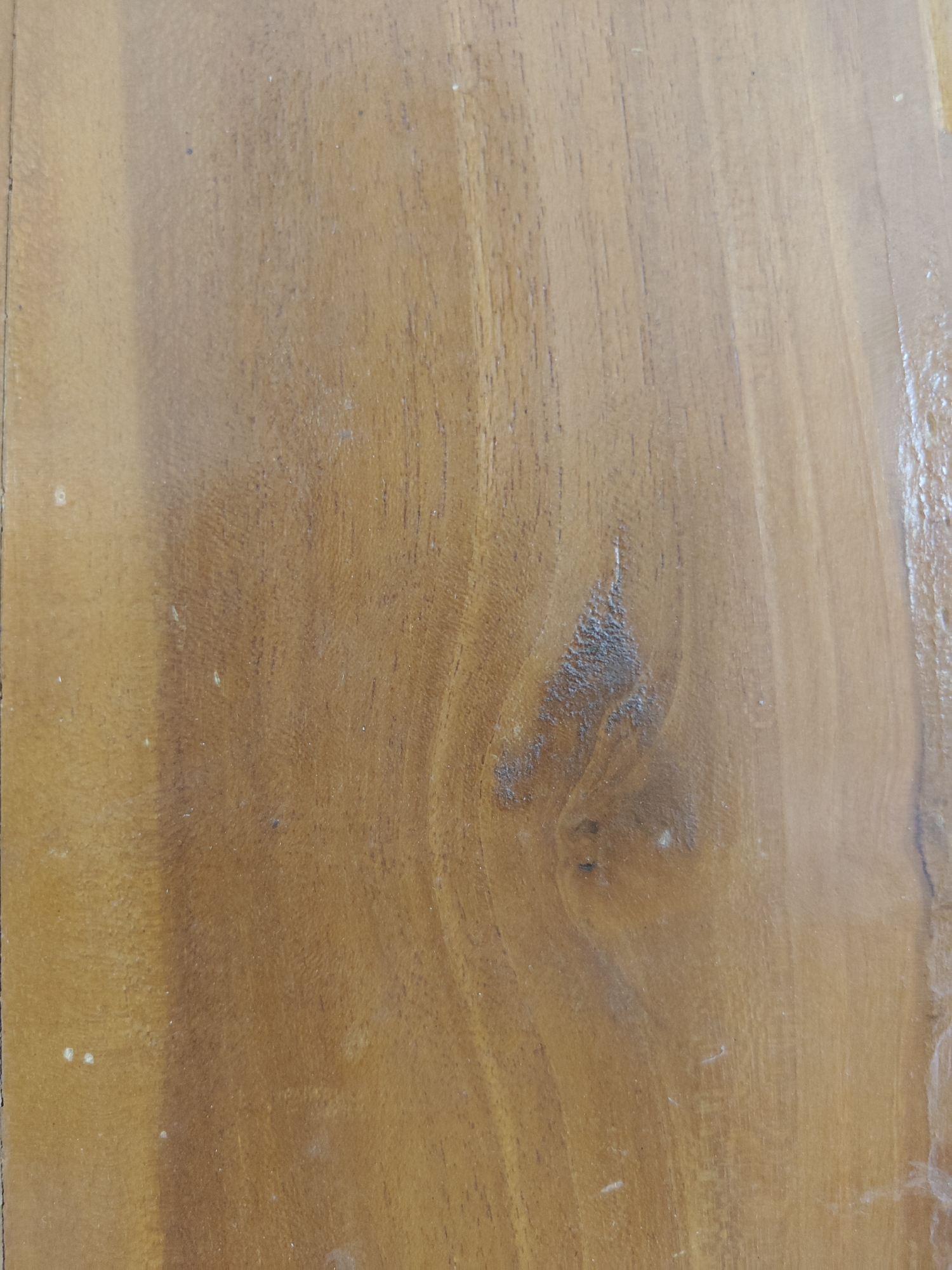 A close-up shot of a polished wooden surface with a natural grain pattern. The wood is a warm, medium-brown color with darker streaks and lighter areas, suggesting variations in the grain. There are a few small white specks scattered across the surface, likely dust or minor imperfections. The texture appears smooth due to the polish. The image is taken in a way that the wood grain runs vertically.