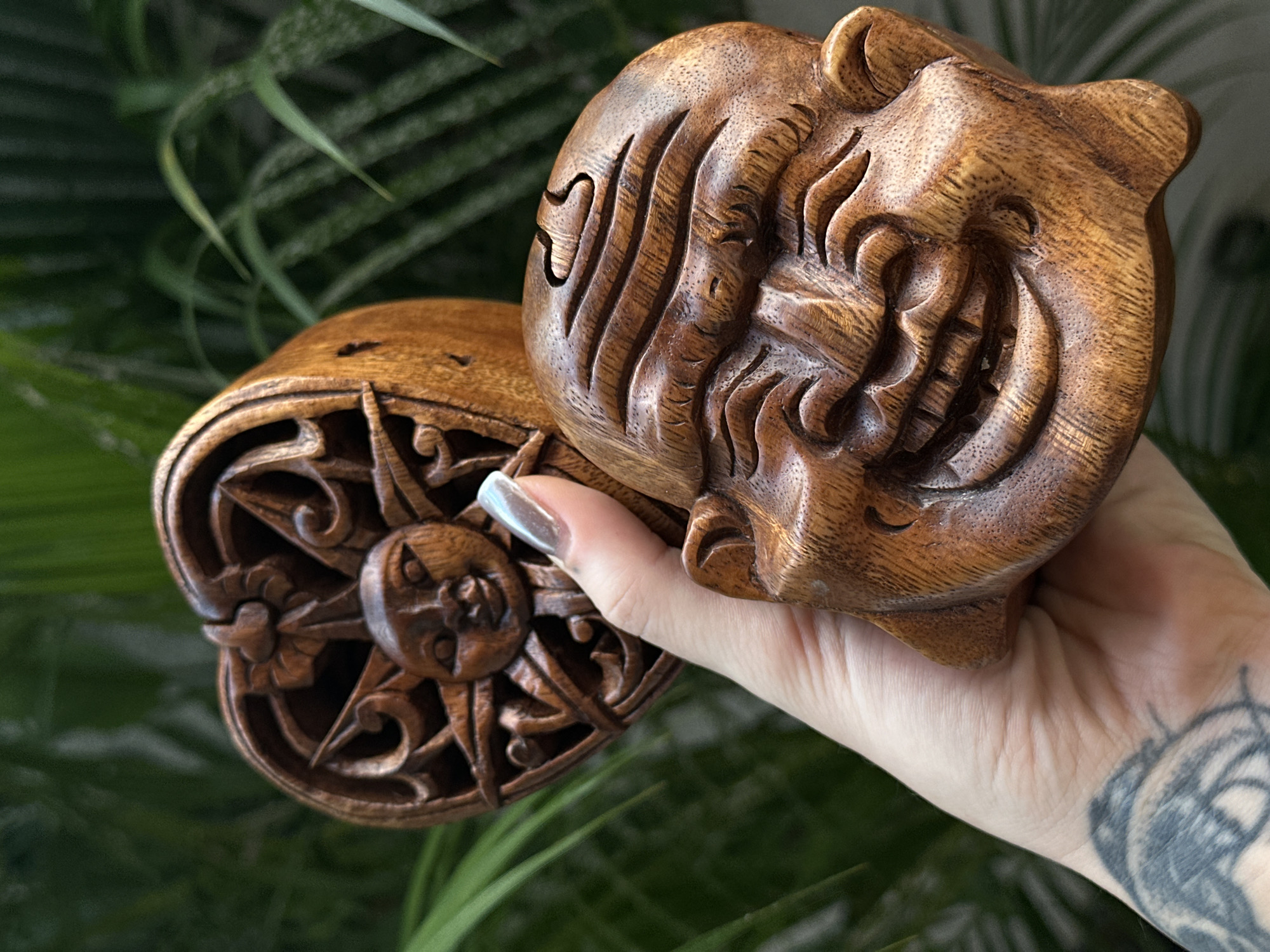 A close-up image showcases a person's hand holding two intricately carved wooden objects against a soft-focus background of lush green foliage, likely captured in North Kuta, Indonesia.
The primary object is a stylized wooden carving of a face, possibly feline or a mythical creature, with deep, flowing lines suggesting fur or facial contours. It is made from a rich, medium-brown wood, highlighting its natural grain and polished surface. This piece is held firmly in the person's right hand.
Partially visible below and to the left of the main carving is another wooden object, featuring a circular design. This piece displays a central carving of a sun or moon face, encircled by elaborate scrollwork and stylized rays. Both objects share a similar warm wooden tone and intricate craftsmanship, suggesting they may be a set or companion pieces.
The hand holding the carvings has fair skin, with a notable tattoo on the wrist featuring radial or ornamental patterns. The thumb displays a long, manicured nail with a subtle iridescent or pearlescent finish.
The background is a vibrant, blurred expanse of green leaves and plant stems, creating a natural and serene setting. The soft, even lighting suggests it is daytime. There is no visible text in the image. The scene emphasizes the detailed artistry of the wooden carvings within a natural context.