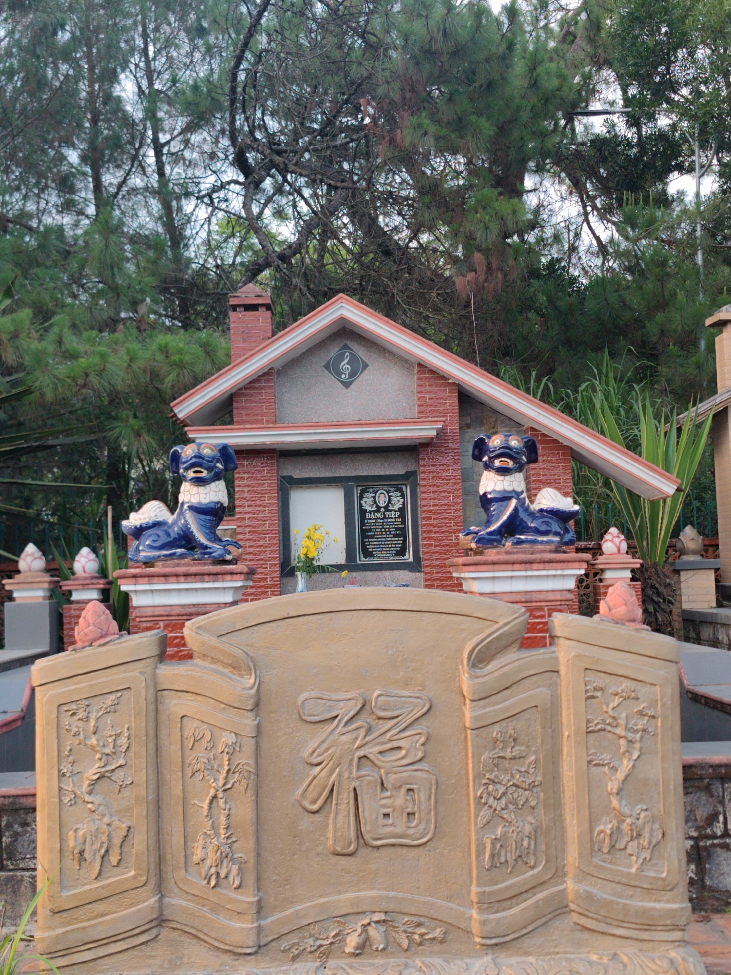 The image depicts a tomb in Cam Ly - Đà Lạt, Vietnam. The tomb features a red-brick structure with a sloping roof, a chimney, and a trefoil clef symbol. Two blue and white lion statues flank a central marker. A floral arrangement sits in front of a smaller inscription. The foreground consists of a tan stone barrier, adorned with carved trees and a large Chinese character. Trees surround the tomb.