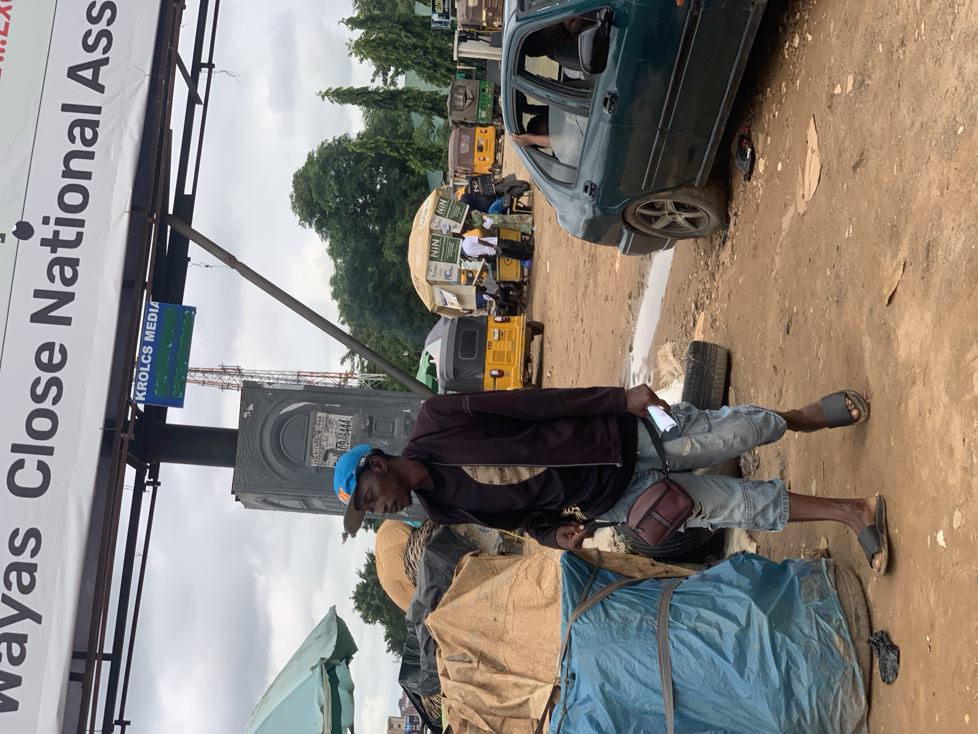 A man wearing a blue baseball cap, a dark jacket over a black t-shirt, and ripped denim shorts walks along a dirt road in a market area. He has a brown satchel slung across his chest and is holding a white document in his right hand. Behind him, to the left, are large piles of goods covered with tarps and a blue plastic sheet. In the background, several yellow and green auto-rickshaws are parked, and a large yellow umbrella with advertisements for "NIN" is set up with people gathered around it. To the right, a dark green car is parked on the side of the road. Above the scene, a large banner with black lettering reads "Wayas Close National Ass". The sky is overcast. The image appears to be taken in Abuja, Nigeria, likely in an outdoor market or transportation hub.