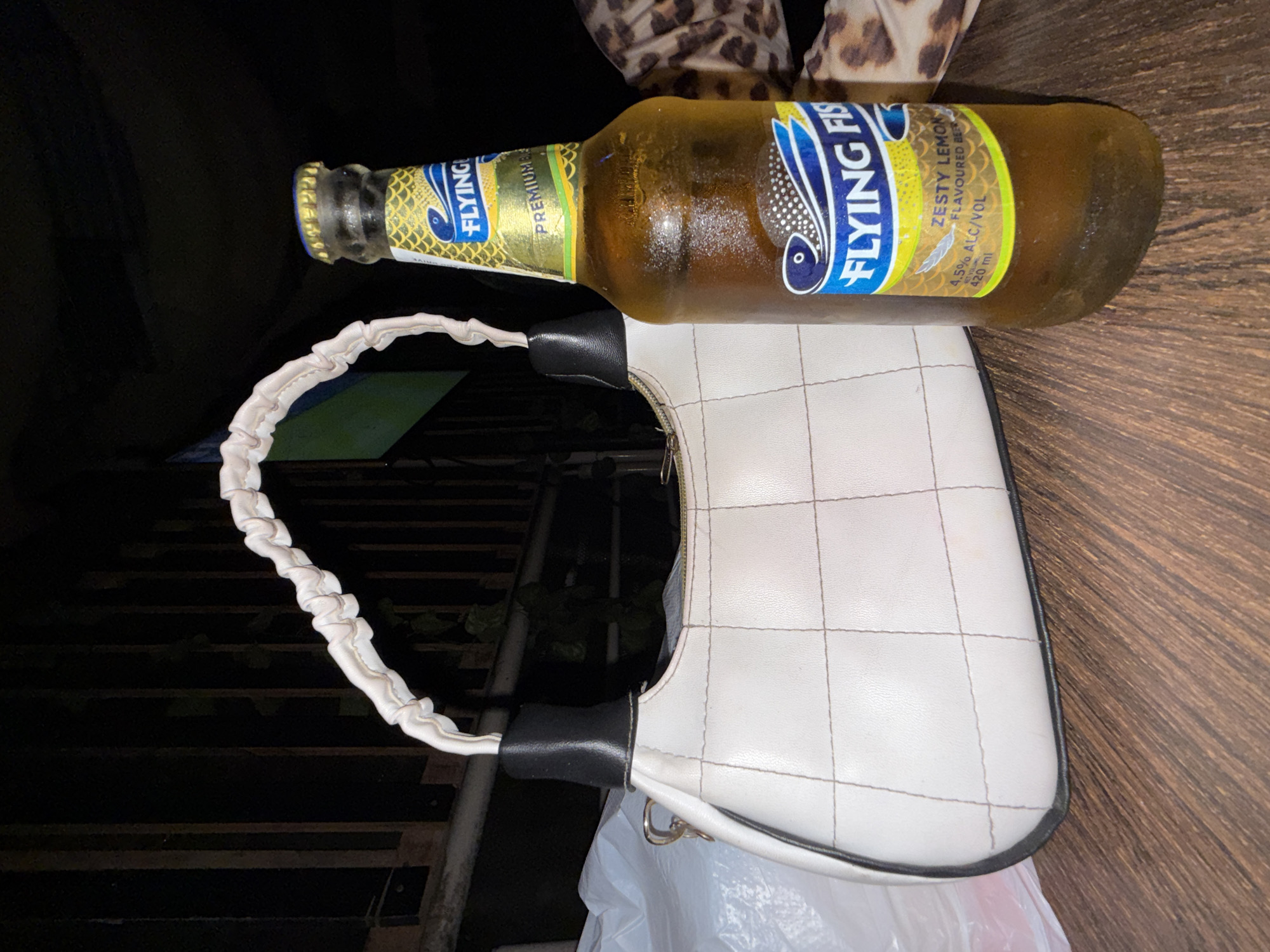 The image shows a bottle of Flying Fish Zesty Lemon flavored beer lying on top of a white quilted handbag with a twisted handle. Part of a leopard print fabric is visible to the upper right of the beer bottle. The background is dark and appears to include a set of stairs. The location in the city of Ado, Nigeria cannot be determined from the image.