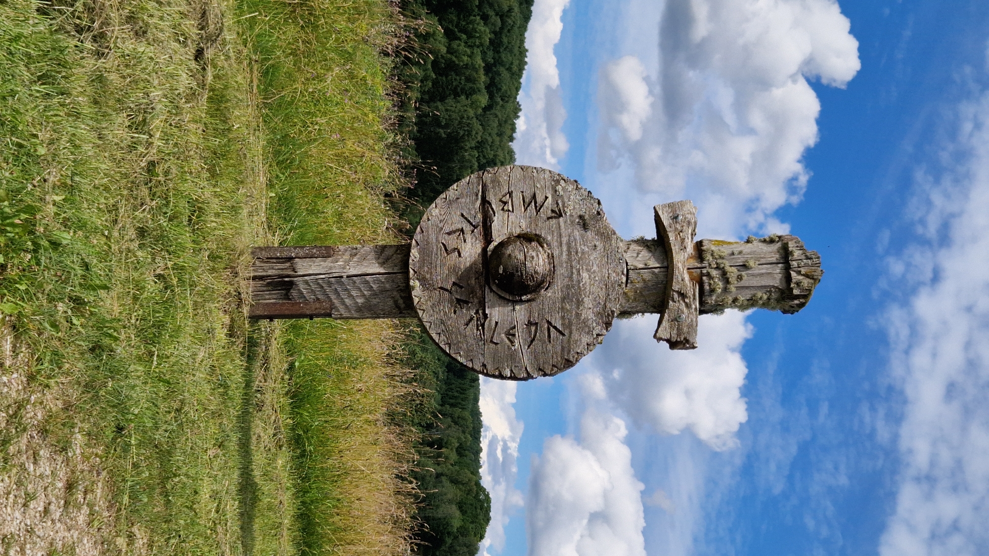 This media file features a wooden cross adorned with carvings, standing in a grassy field under a bright blue sky with fluffy white clouds. The cross is made of rough-hewn wood, weathered and covered in patches of lichen. A circular wooden disc is attached to the vertical beam of the cross, and this disc also bears carved symbols.
Visible text on the wooden disc appears to be in a runic script. One segment of the inscription is clearly legible and reads: "MBVTS". Below this, another partially visible inscription seems to say "NLAGN". The overall scene suggests an outdoor location, possibly in a rural or historical setting. The lighting indicates it is daytime, with the sun casting shadows. The sky is clear and sunny, suggesting fair weather. The background shows a dense line of green trees, possibly a forest, and a sloping field of dry grass and wildflowers. The composition is vertical, with the cross dominating the center of the frame.