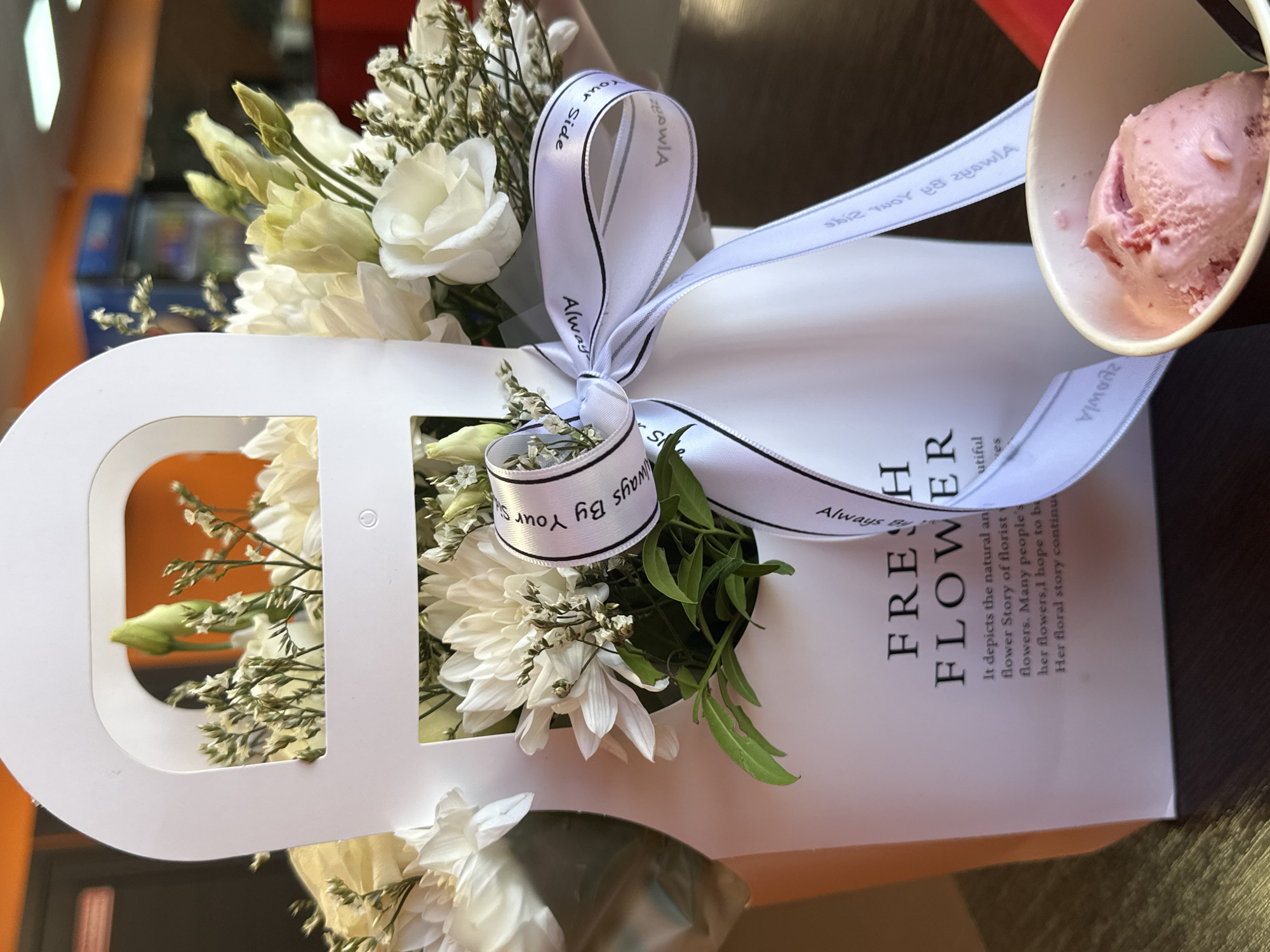 A high-angle shot shows a bouquet of white flowers in a white box with "FRESH FLOWER" printed on it, tied with a white ribbon that reads "Always By Your Side," next to a cup of pink ice cream. The bouquet contains a variety of white flowers and greenery. The box has cutout sections revealing the flowers inside. The ice cream is in a small, white cup. The scene is lit well with natural light, suggesting an indoor setting. A dark surface can be seen underneath the bouquet and the ice cream.