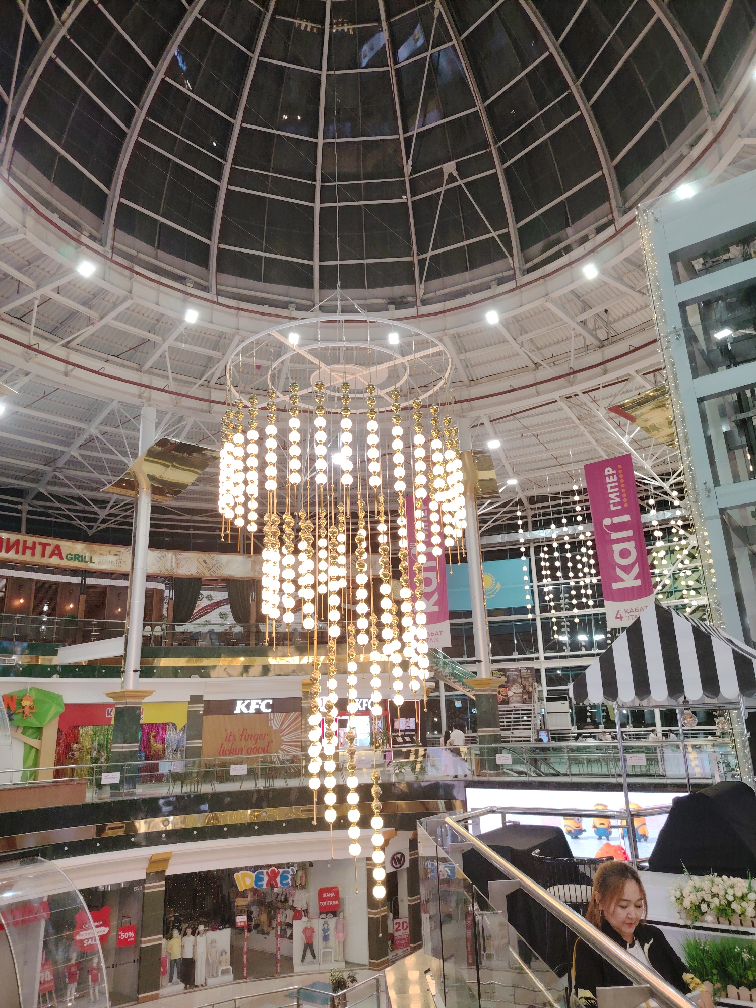 The image depicts the interior of a multi-level shopping mall, identified as Mega Silk Way in Astana, Kazakhstan. A large, ornate chandelier with numerous glowing orbs hangs prominently in the center of the frame, suspended from a circular frame beneath a geodesic glass dome ceiling. Various retail establishments are visible on different levels, including KFC, "kаri" (a footwear store), and "Pinta Grill". Banners and signage in Cyrillic script indicate the location. A woman is seated on a set of stairs on the right side of the image, looking down. Other individuals can be seen in the background, navigating the mall on various levels. The environment is well-lit, with artificial lights supplementing the natural light filtering through the dome.